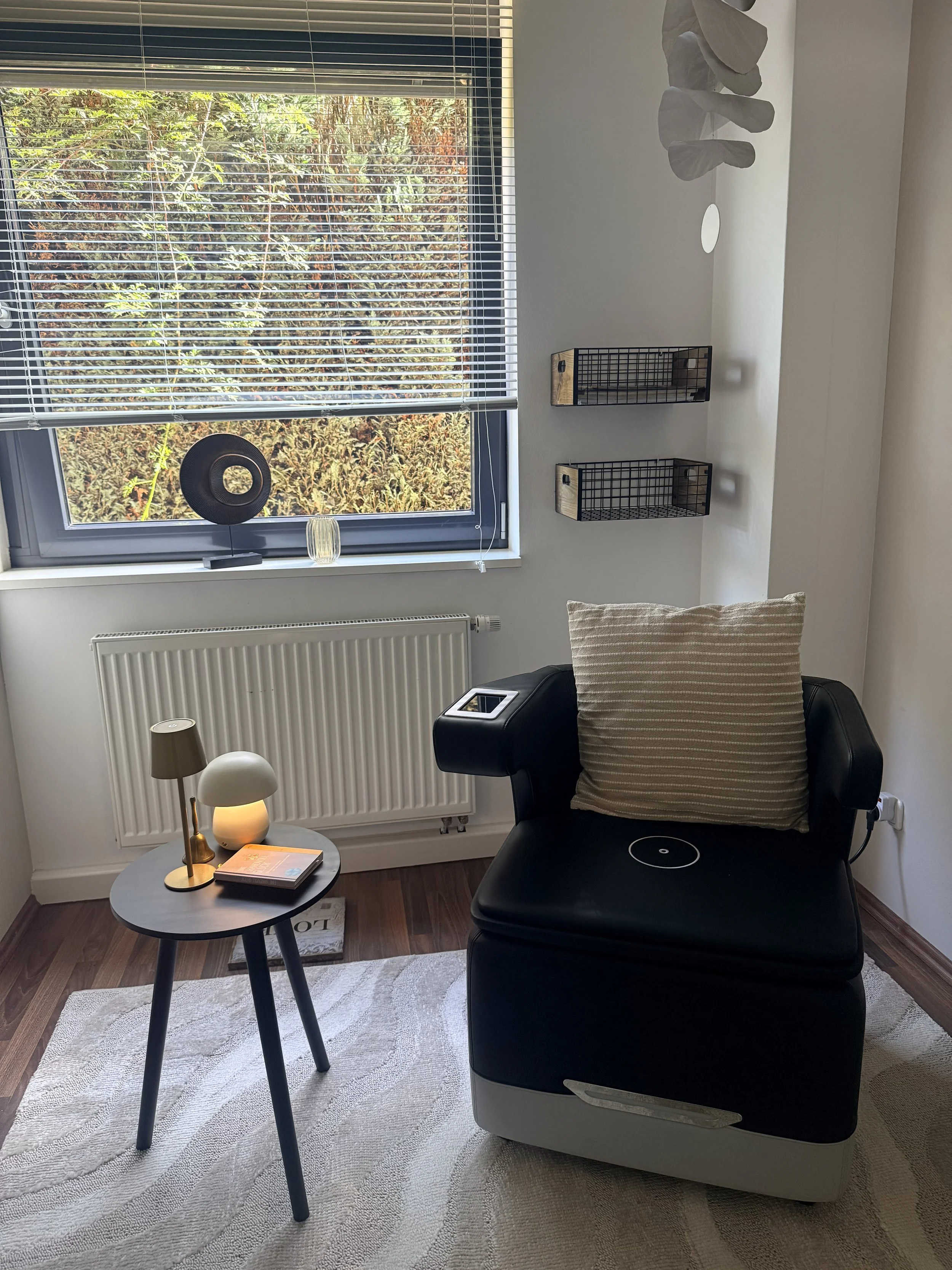 Ein modernes Wohnzimmer mit einem schwarzen Sessel, einem kleinen runden Tisch mit Büchern, einer Stehlampe und einem Fenster mit offenen weißen Rollos. An der Wand hängen zwei schwarze Wandregale, eine Skulptur aus weißem Stein oder Keramik und eine weiße Vase.