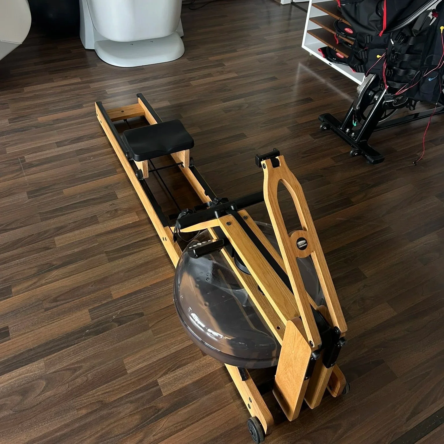 Ein Holzrollator mit schwarzen Sitz und einem transparenten Regenhaube, auf einem Holzboden, im Hintergrund ein WC und eine Hygiene-Station
