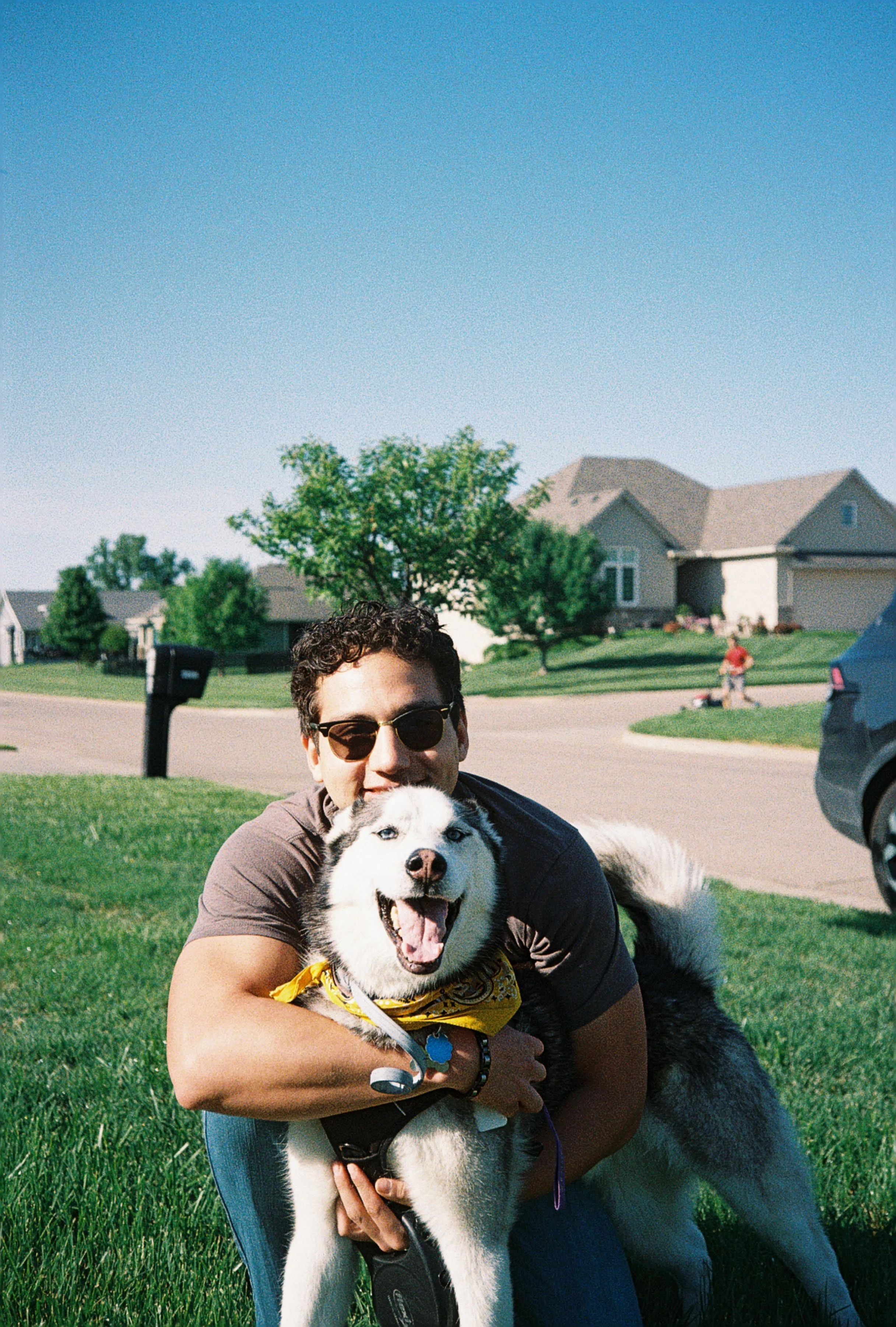 A man with dark, curly hair and sunglasses hugging a happy Siberian Husky dog on green grass in a suburban neighborhood with houses, trees, and a person walking in the background during daytime.