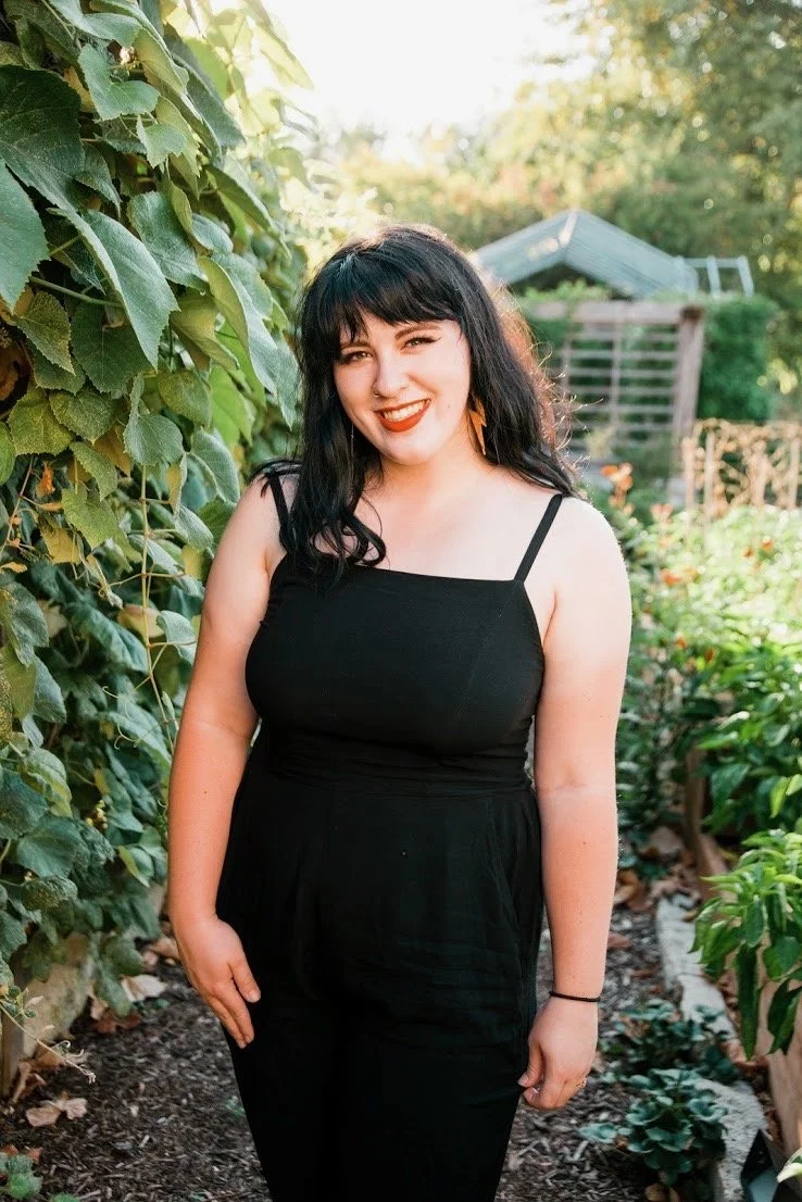 A woman with black hair, wearing a black sleeveless top, standing outdoors in a garden, smiling and winking.