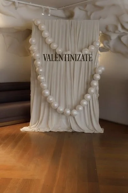 Decorative display with a draped white fabric backdrop, large pearl garland, and the word 'VALENTINAZATE' in black letters, set on a wooden floor.