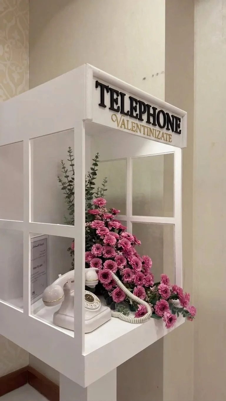 A vintage white telephone with a rotary dial on a decorative white stand decorated with pink flowers and green foliage, with a sign reading 'TELÉPHONE VALENTINIZATE' on top.
