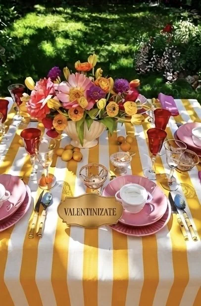 Spring outdoor table setting with a yellow and white striped tablecloth, pink plates, gold flatware, colored glasses, and a large colorful flower arrangement as the centerpiece, with a sign reading "Valentinizate" in front.
