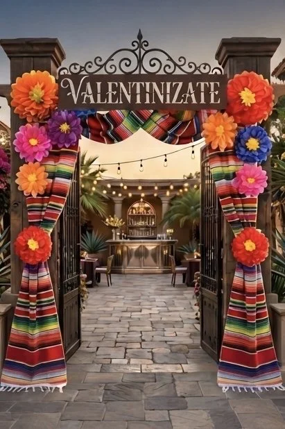 Decorated entrance with a sign that says 'Valentinízate', floral paper flowers, colorful striped Mexican blankets, and string lights, leading to an outdoor patio area.