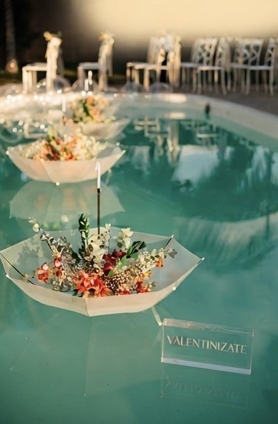Floating floral arrangements on a pool, with a sign reading 'Valentinzate' nearby, and chairs in the background for a celebration or event.