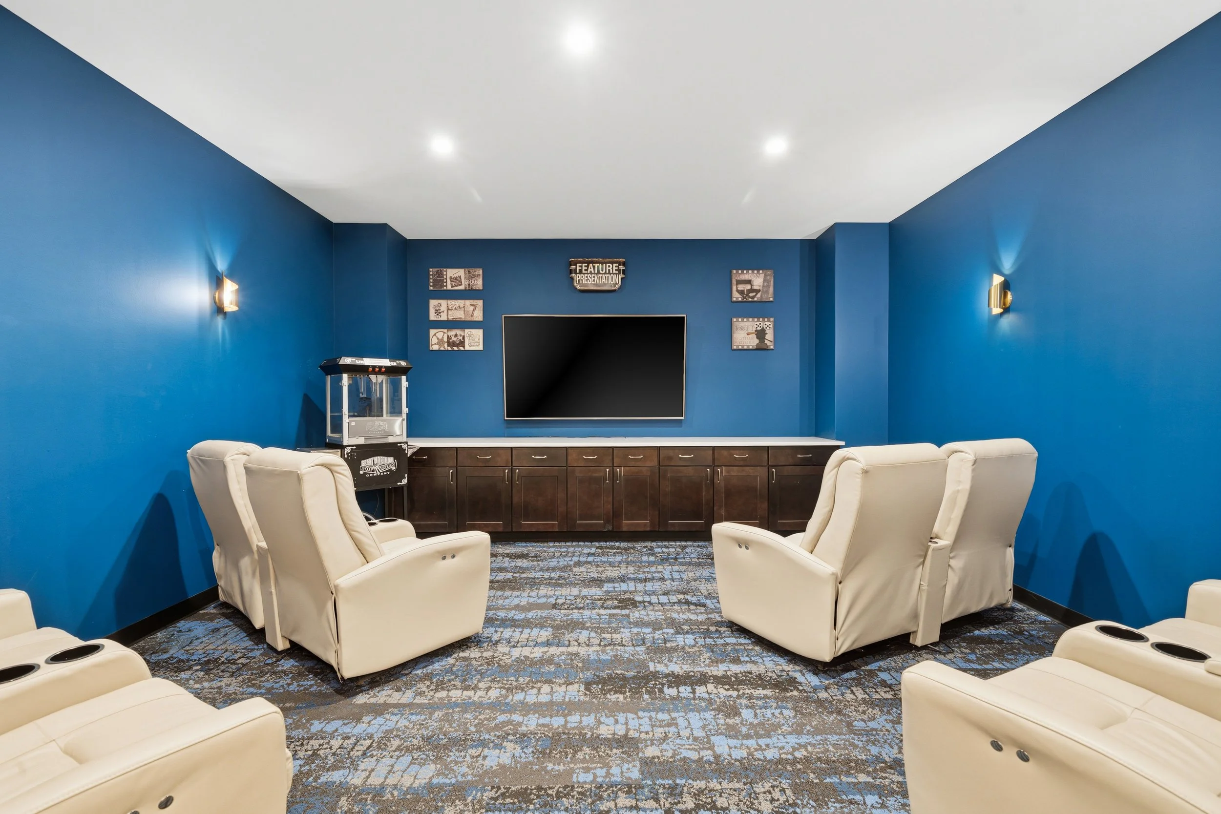 A modern conference room with blue walls and white chairs arranged in a semi-circle facing a large flat-screen TV on a wooden cabinet. The room has decorative wall art and wall-mounted lights.
