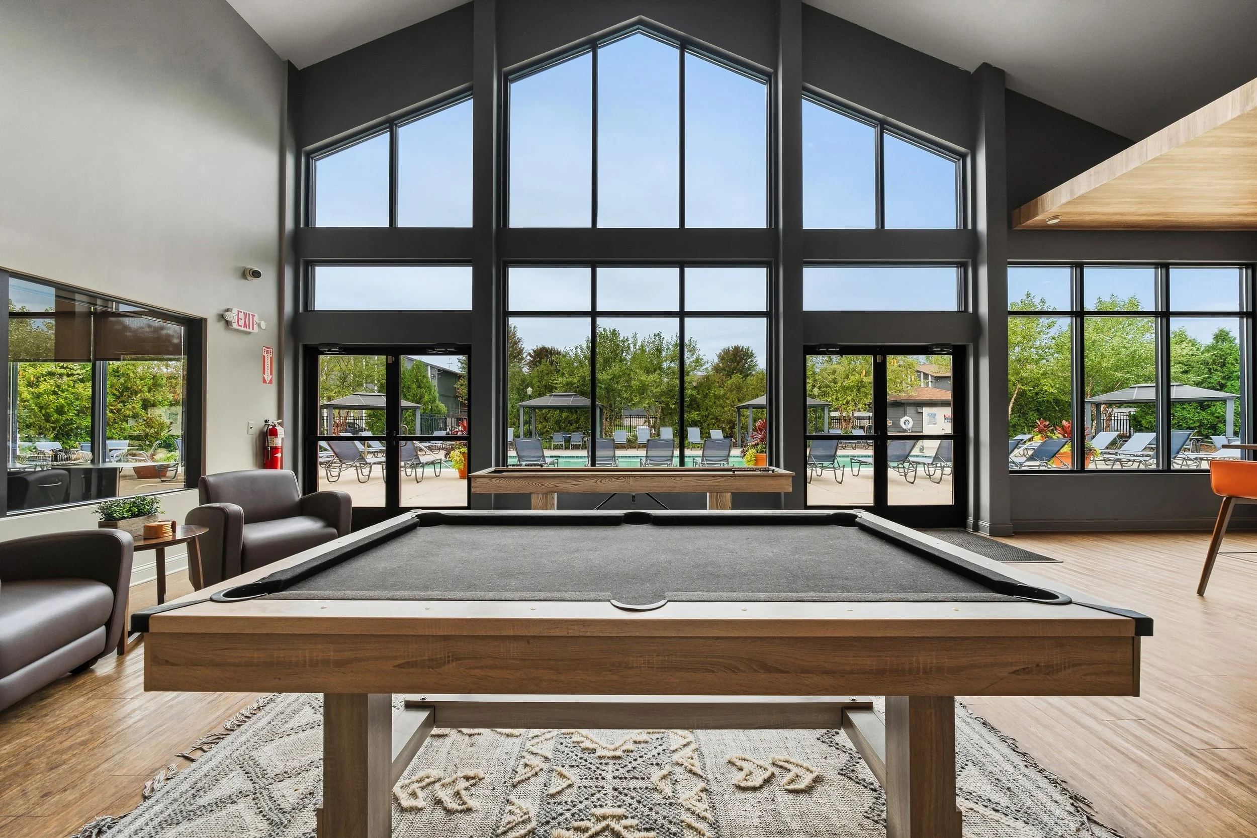 Interior view of a recreation room with a pool table, chairs, and large windows overlooking a pool area with lounge chairs and green trees.