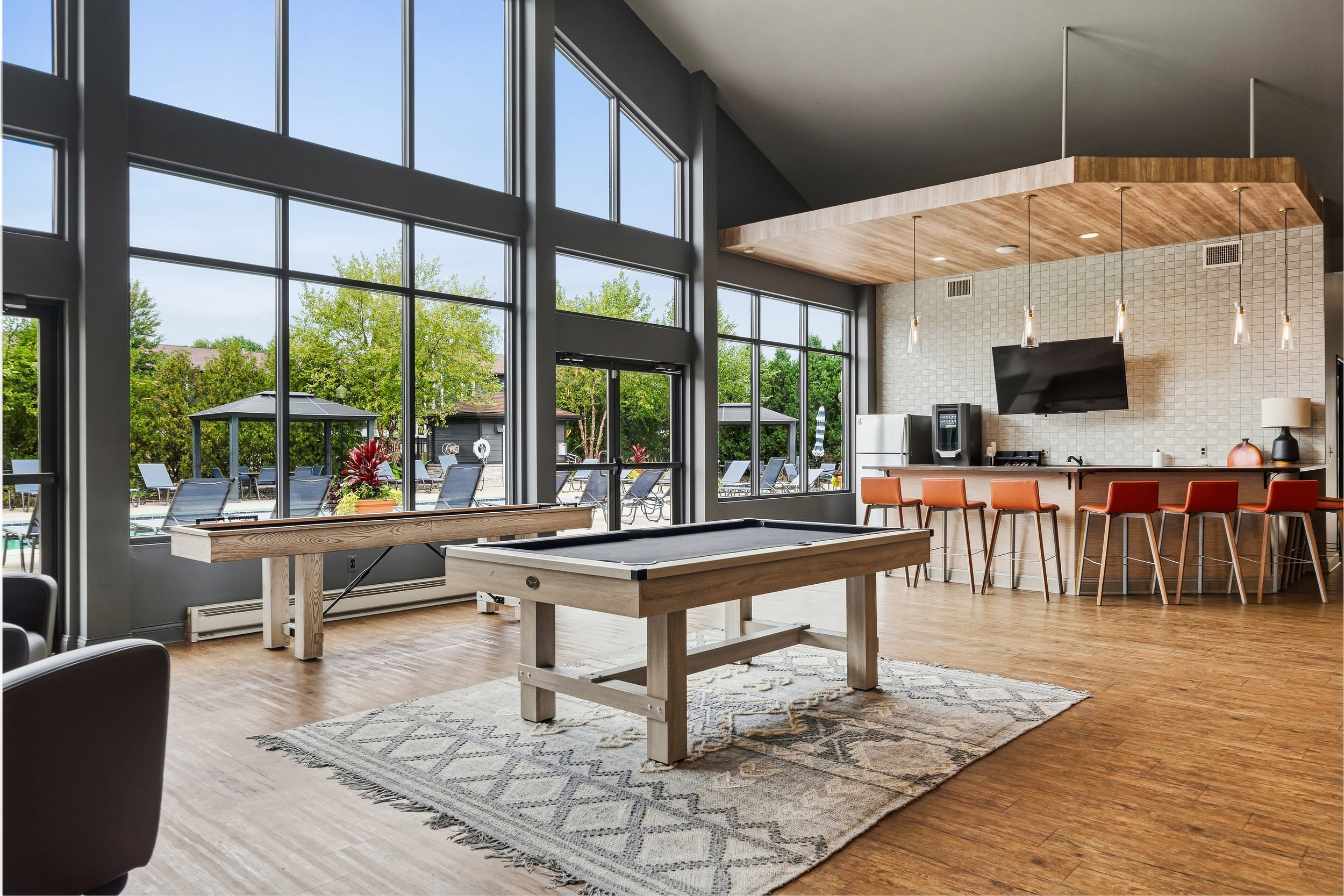 Interior of a modern community room with large windows, a high ceiling with wooden accents, and a bar area with orange stools. The room includes a foosball table, a patterned rug, and outside there's a patio with lounge chairs and an umbrella.