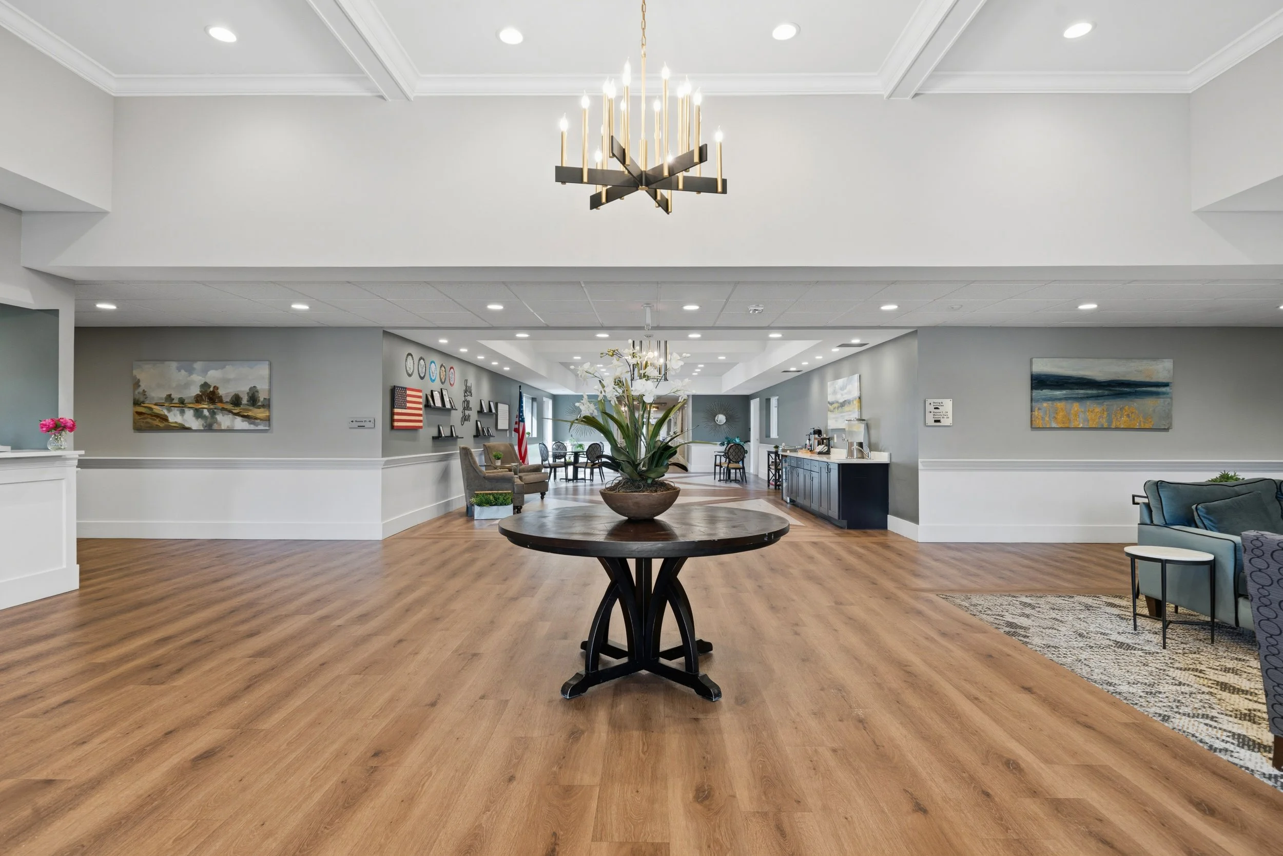 Interior of a spacious, modern lobby or common area with a wooden floor, a round table with a floral arrangement, decorative paintings on the walls, a chandelier hanging from the ceiling, and seating areas in the background.