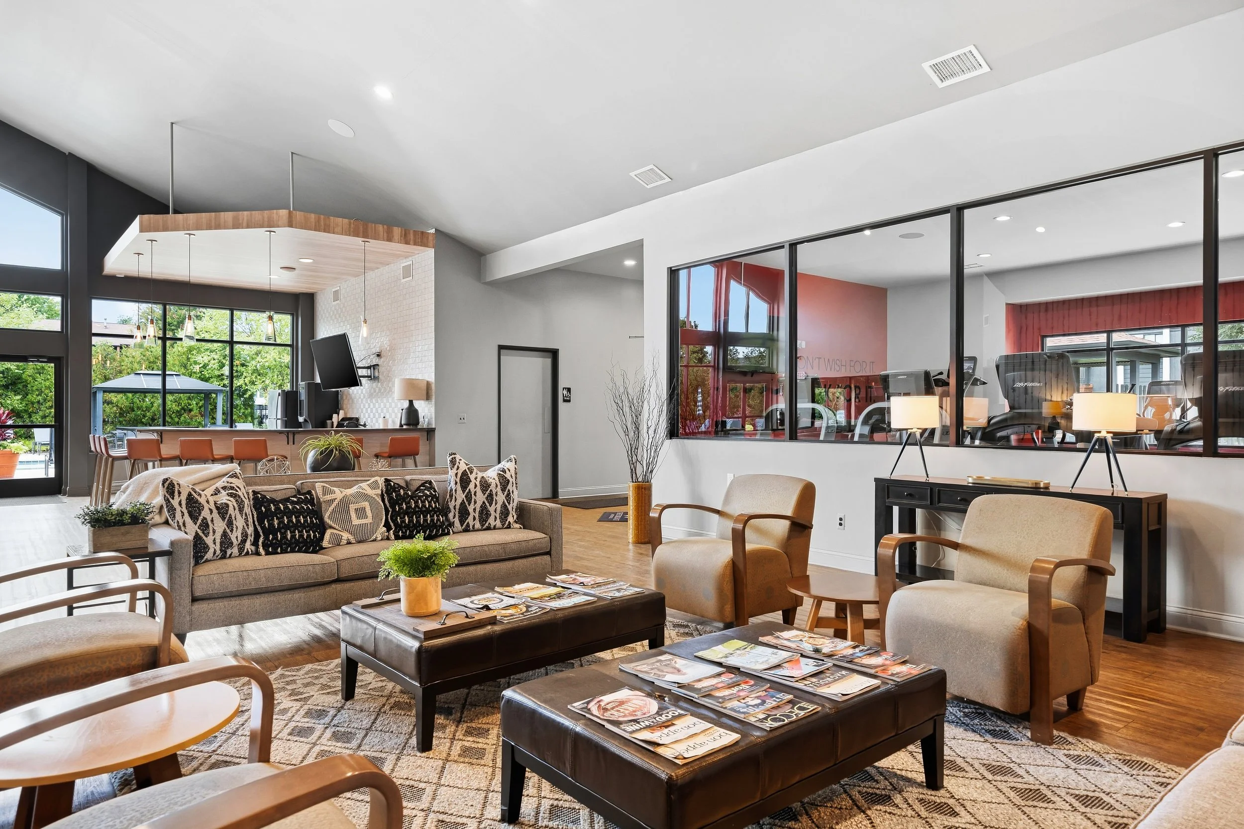 Interior of a modern lounge with large windows, featuring a beige sofa with black and white patterned pillows, wooden chairs, and coffee tables with magazines, decorated with plants, lamps, and a wall-mounted TV.