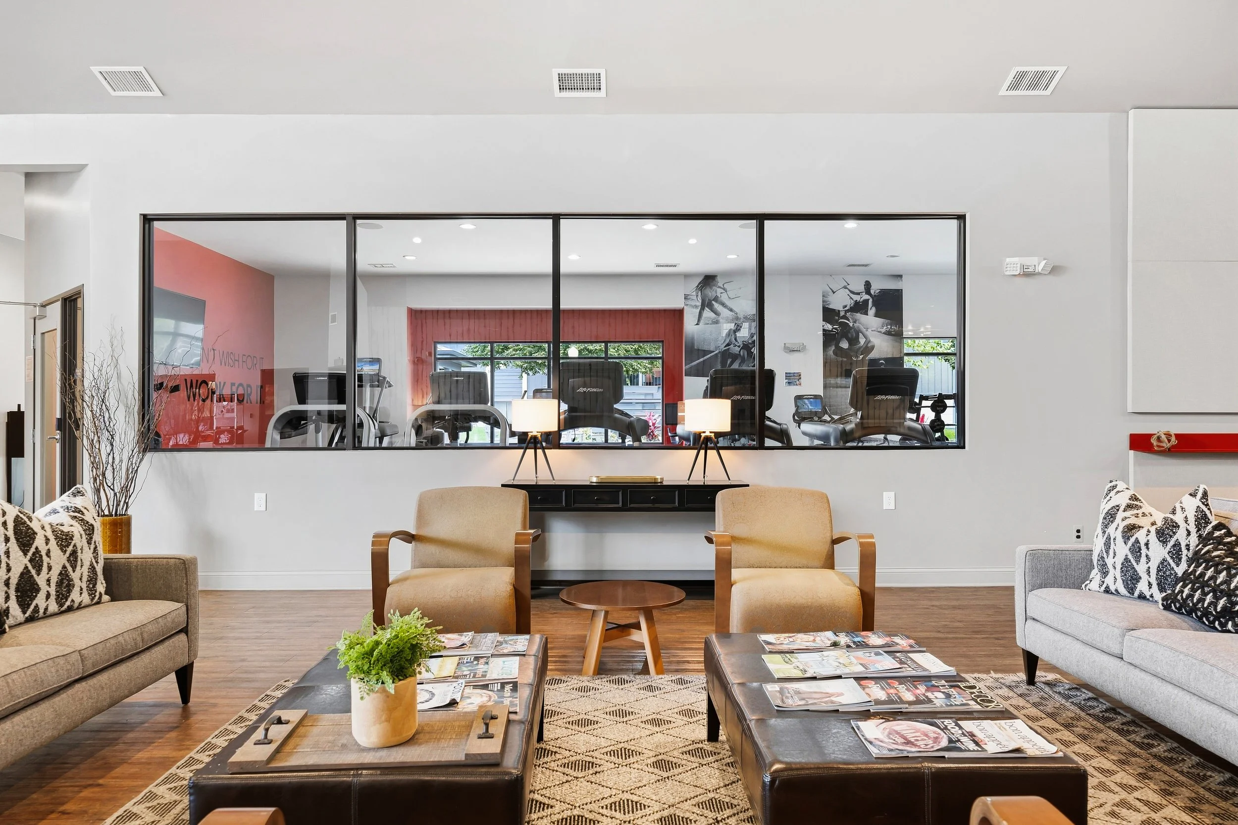 Living room with beige sofas, coffee tables with magazines, potted plant, and view through large glass windows showing exercise equipment in a fitness room with wall art, inside a modern home.