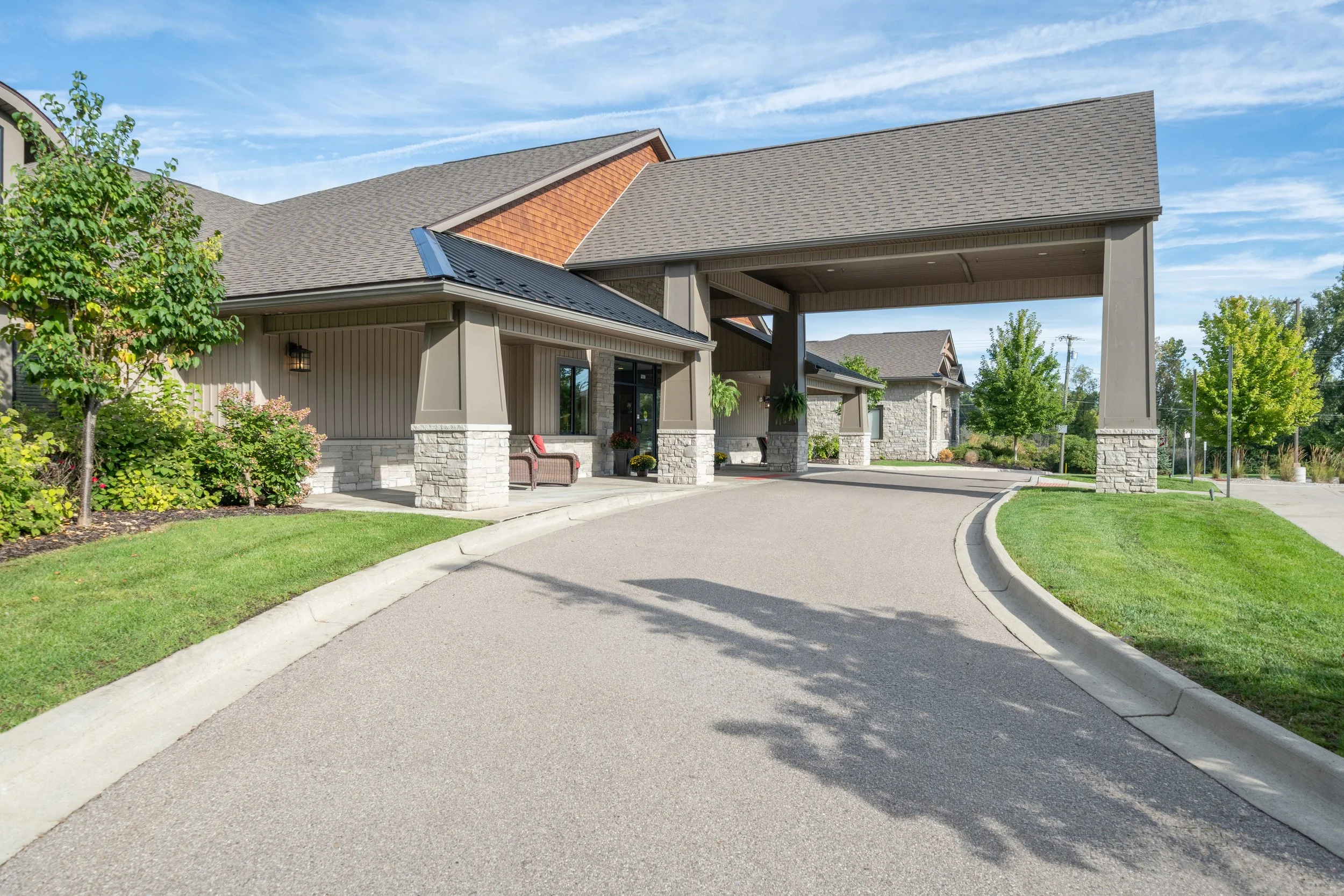 A modern residential building with a covered entrance, stone and siding exterior, and a car driveway, surrounded by greenery and trees under a blue sky.