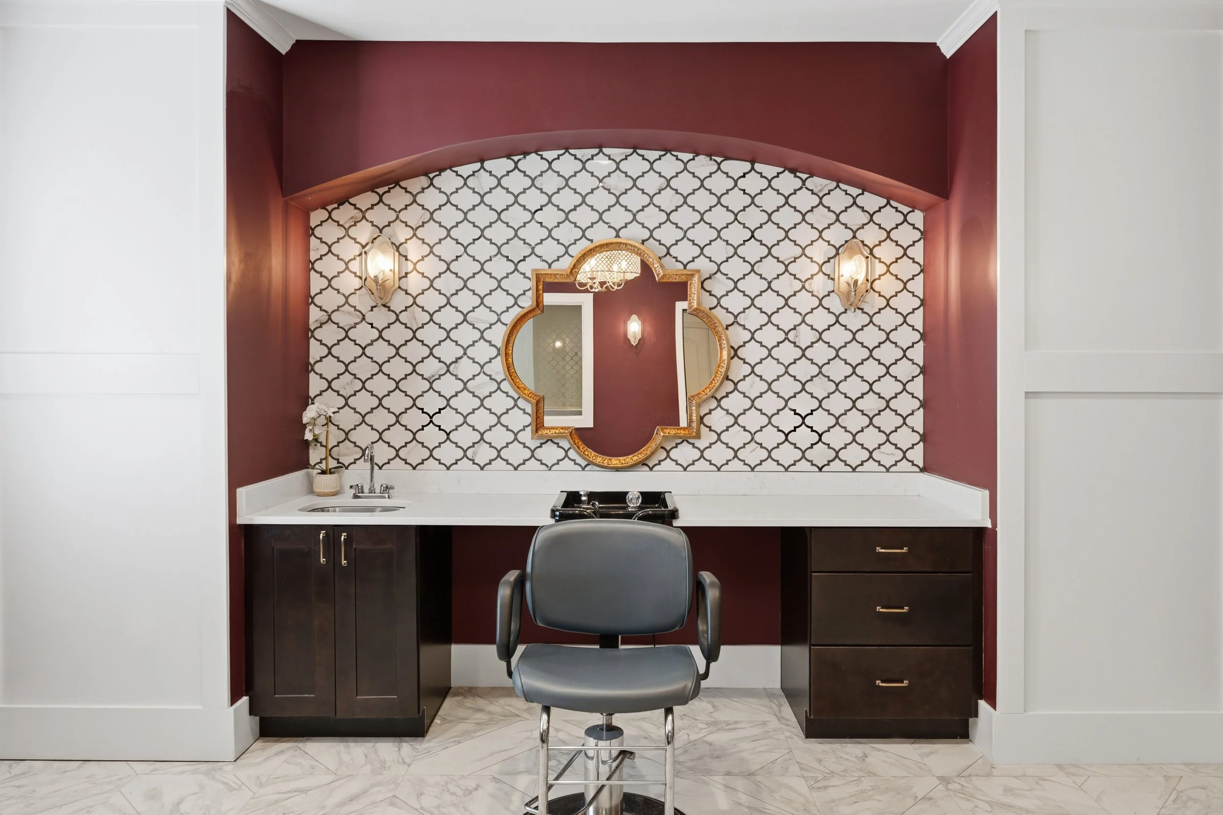 A beauty salon station with a mirror-shaped shelf, surrounded by decorative wallpaper, light fixtures, and a marble countertop with a faucet and a black chair in front.