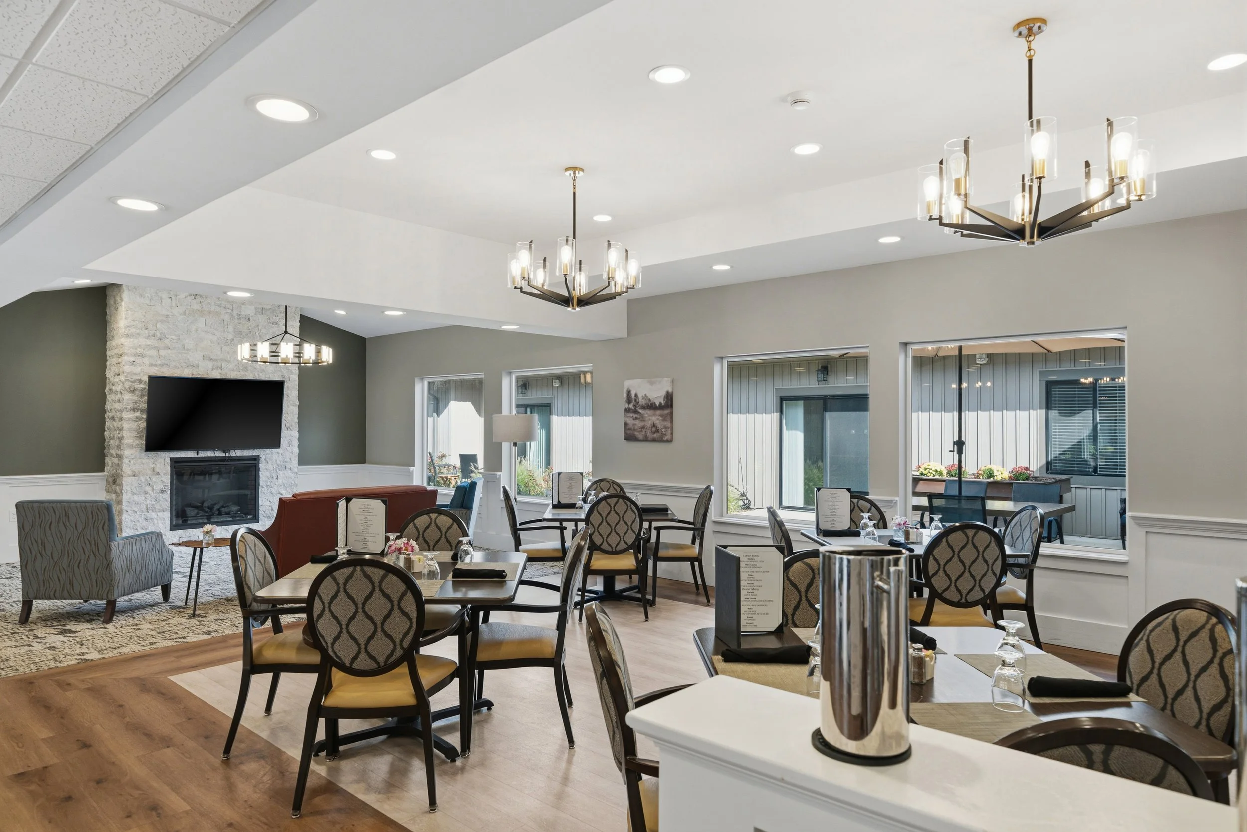Interior view of a modern restaurant with multiple dining tables, patterned chairs, and large windows letting in natural light. Elegant lighting fixtures hang from the ceiling, and a stone fireplace with a television mounted above it is visible in th