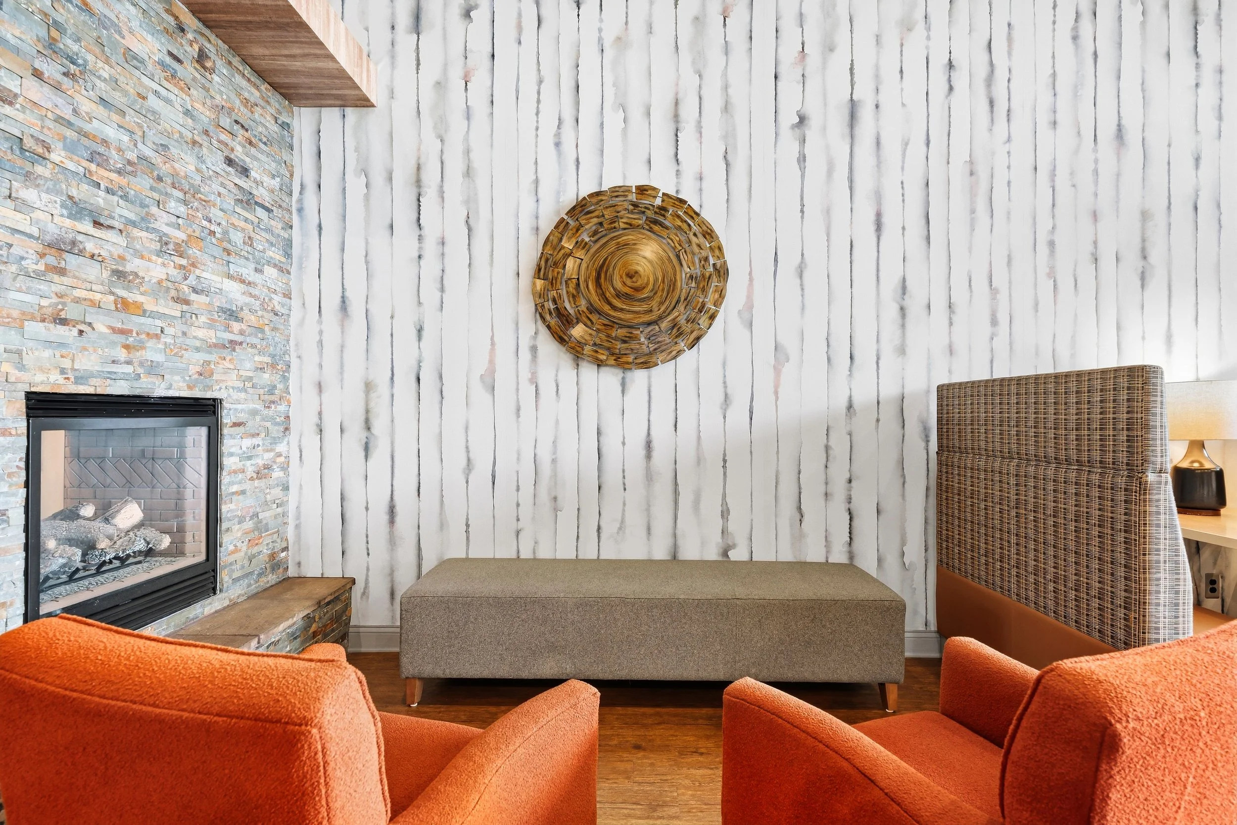 Living room with a stone fireplace, a patterned wall art piece, a bench, a side table with a lamp, and orange chairs surrounding it.