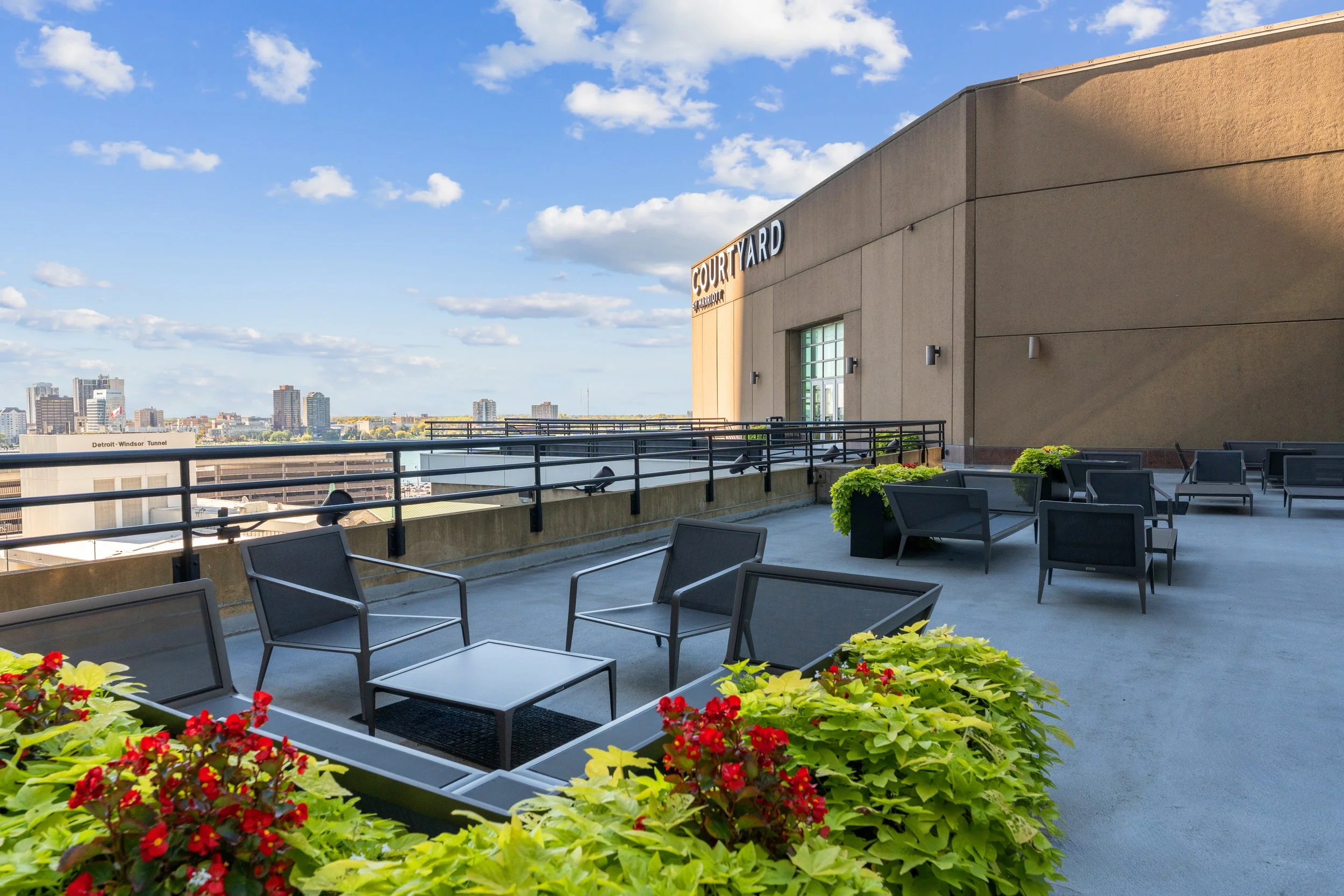 A rooftop terrace with tables, chairs, and planters with red and green flowering plants. In the background, there is a city skyline under a partly cloudy sky. The building has a sign that reads 'Court YARD' and a view labeled 'Detroit - Windsor Tunne