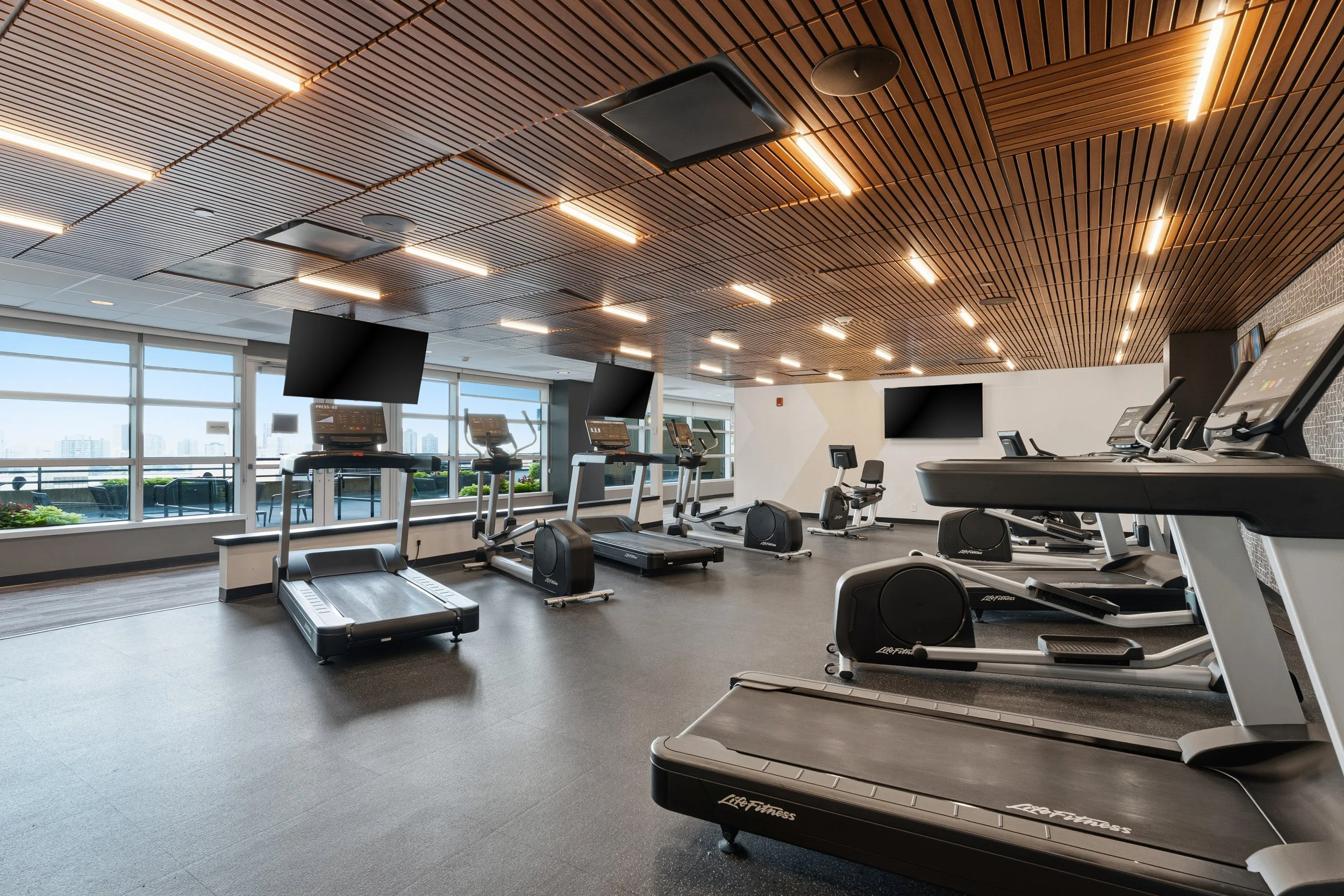 Gym with treadmill and elliptical machines facing large windows, contemporary ceiling design with wooden panels and LED lighting, and flat screen TVs