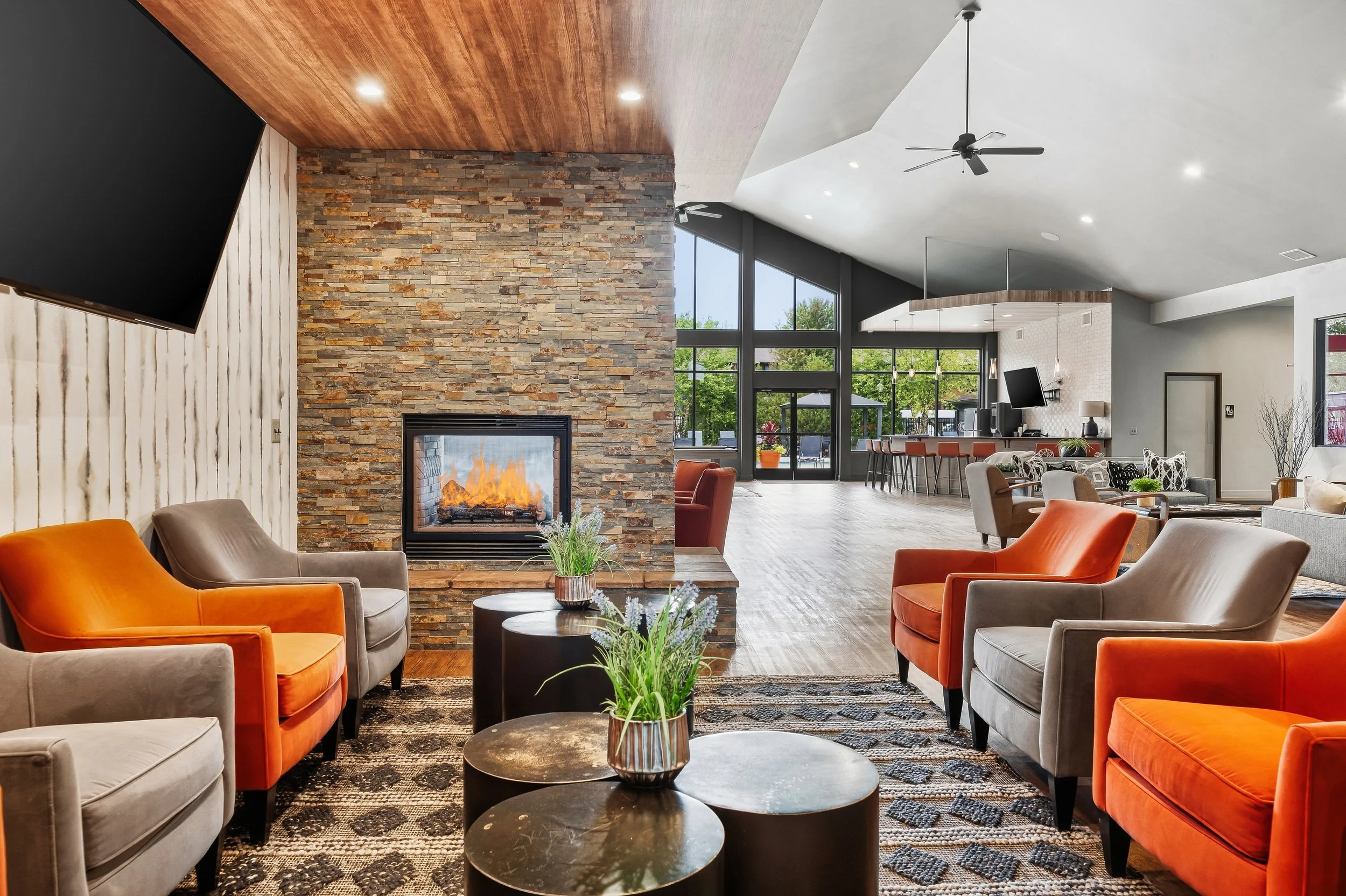 A spacious living room with a stone fireplace, large windows, and modern furniture including orange and gray armchairs, black round coffee tables, and decorative plants.