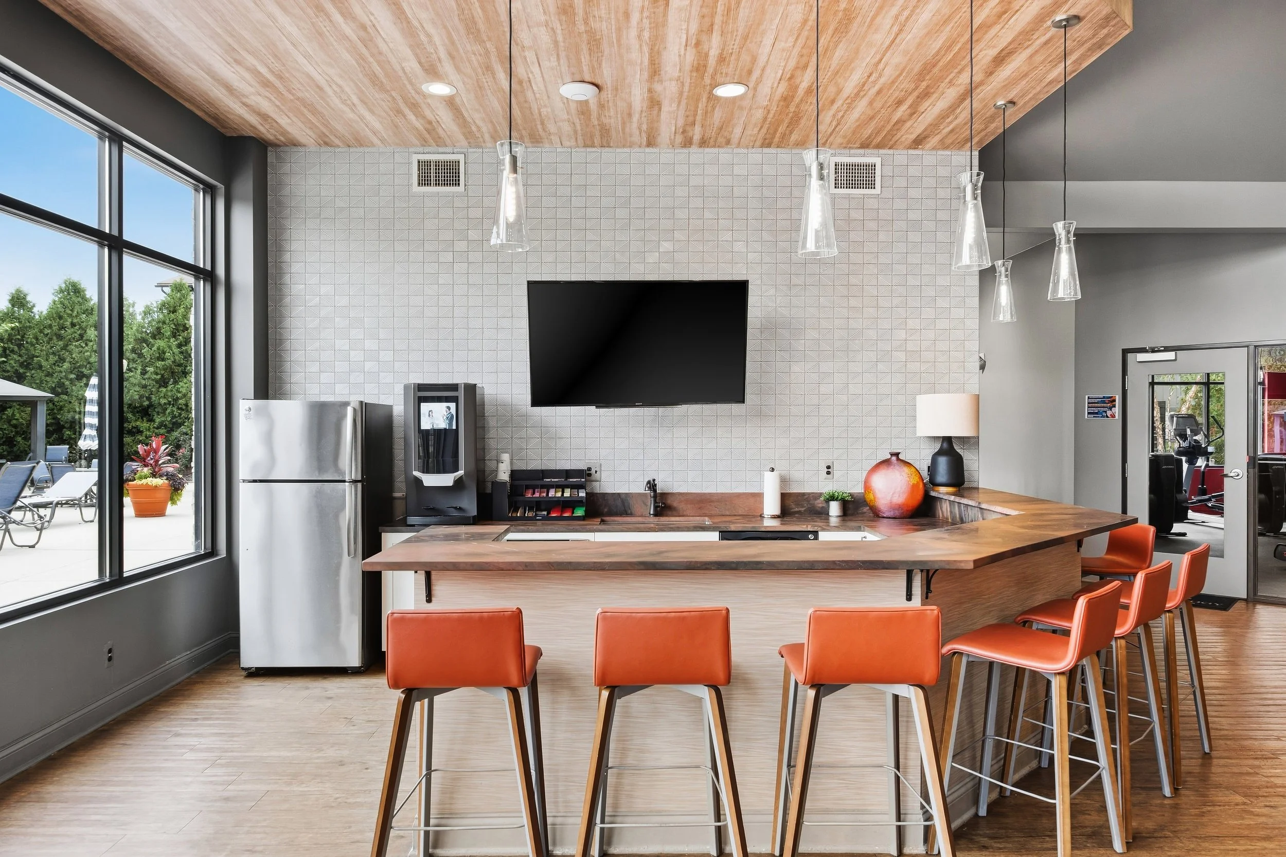 Modern kitchen with a large window, a wall-mounted TV, orange chairs around a wooden island, pendant lights, and a view of an outdoor patio with lounge chairs and trees.
