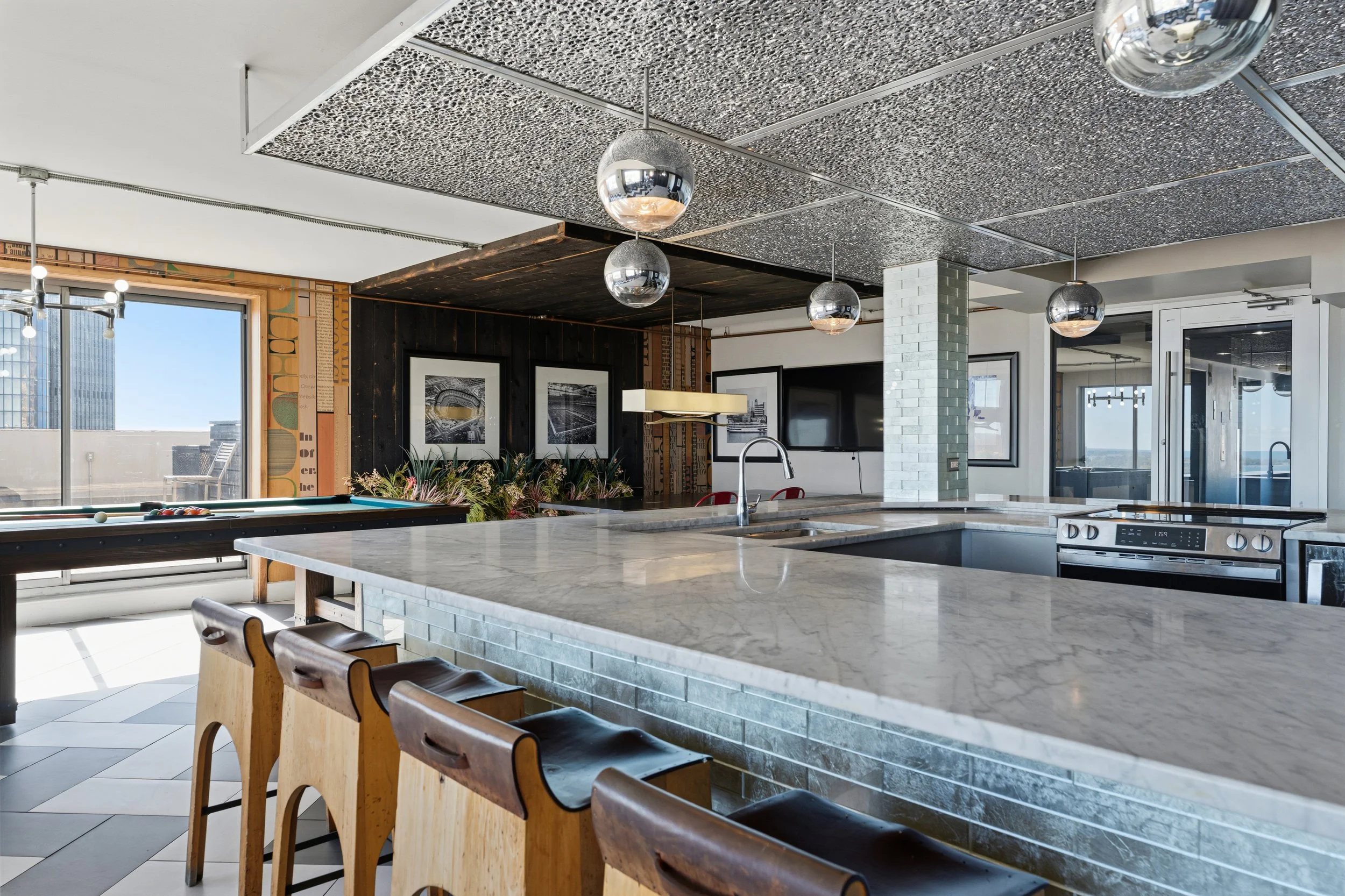 Modern kitchen with a marble island, wooden stools, pendant lighting, large window, and cityscape view.