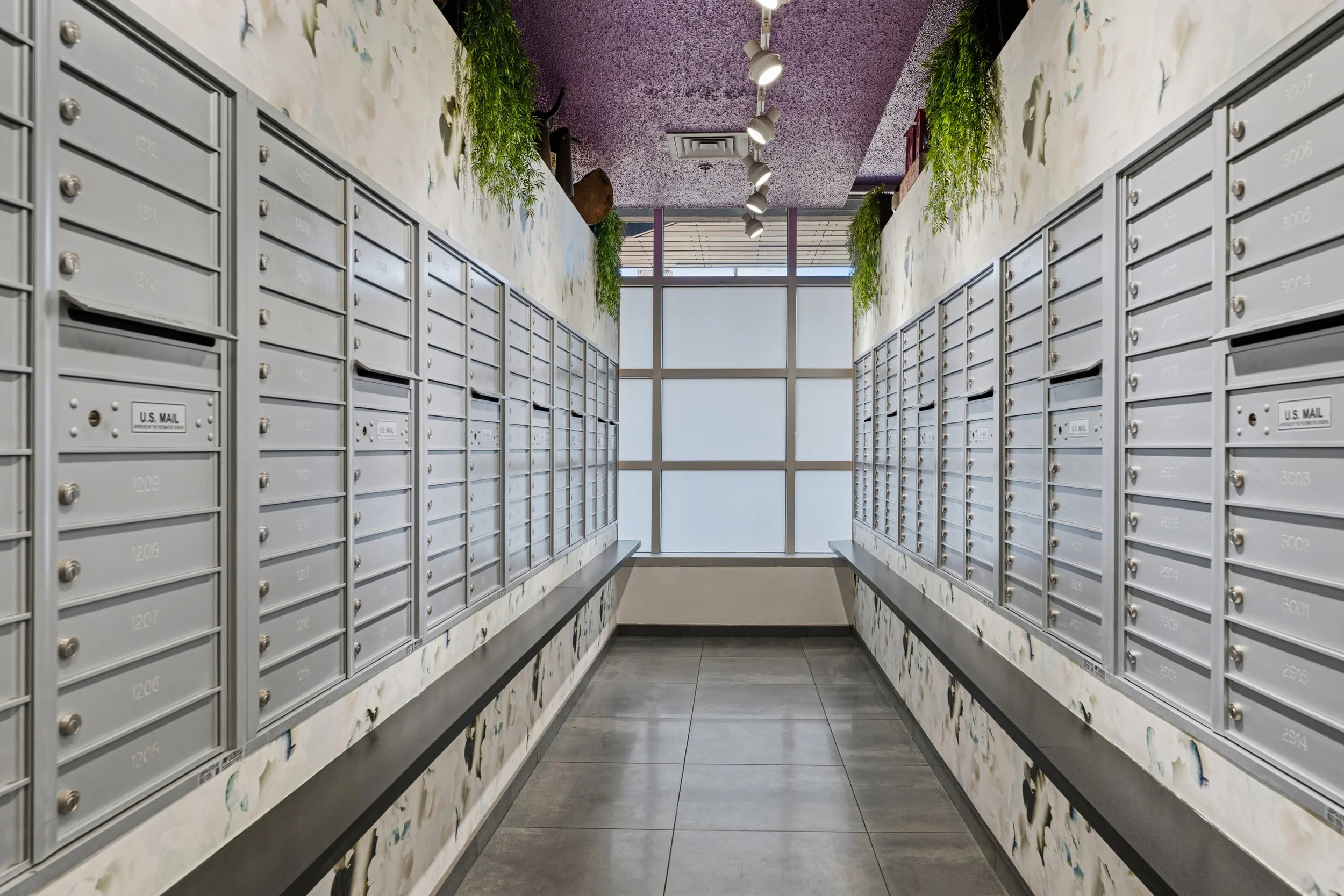 Indoor mailroom with two parallel walls of gray mailboxes with silver locks, decorated with faux marble and floral wallpaper, green hanging plants, and a large window with blinds at the end.