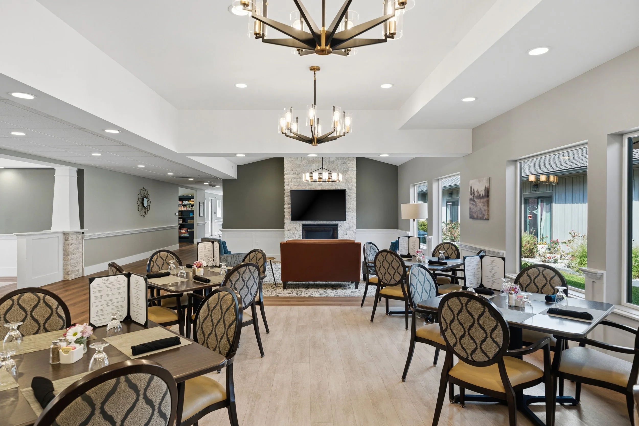 Interior of a restaurant or cafe with tables set for dining, including floral centerpieces, menus, glasses, and utensils. The room has large windows, modern chandeliers, a stone fireplace with a television above, and neutral-colored walls.