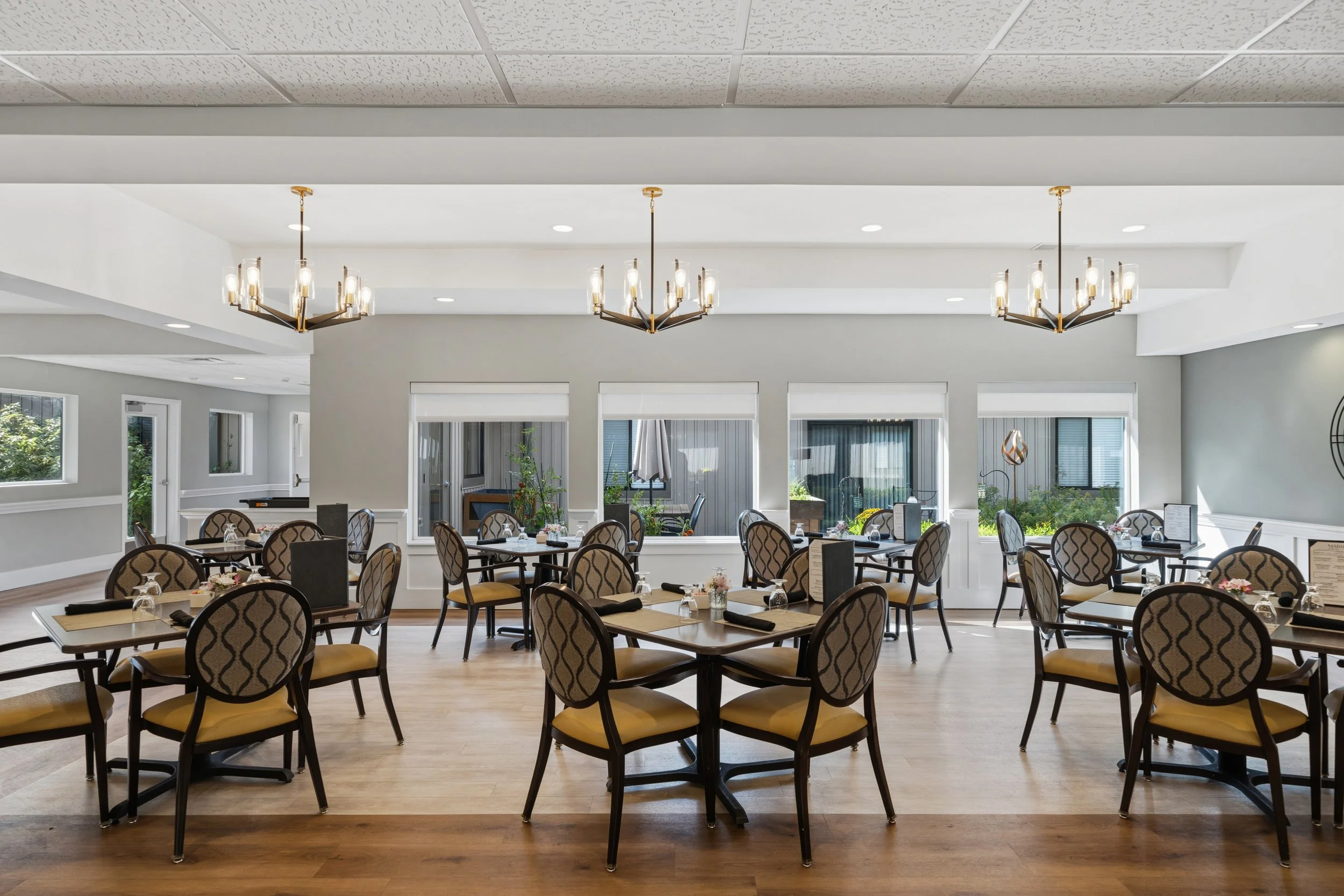 A spacious dining area with multiple rectangular tables, elegant chairs with patterned upholstery, large windows, and hanging chandeliers with multiple bulbs.