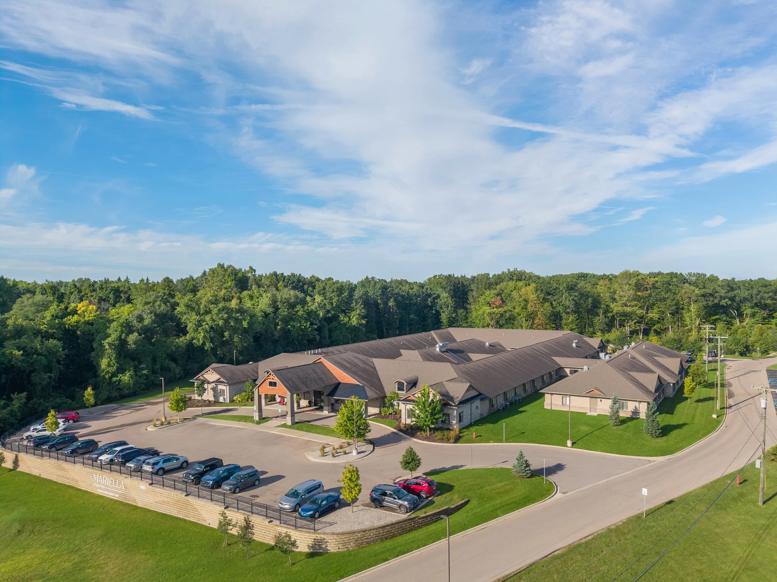 Aerial view of a residential neighborhood with houses, trees, a parking lot with cars, and a clear blue sky with some scattered clouds.