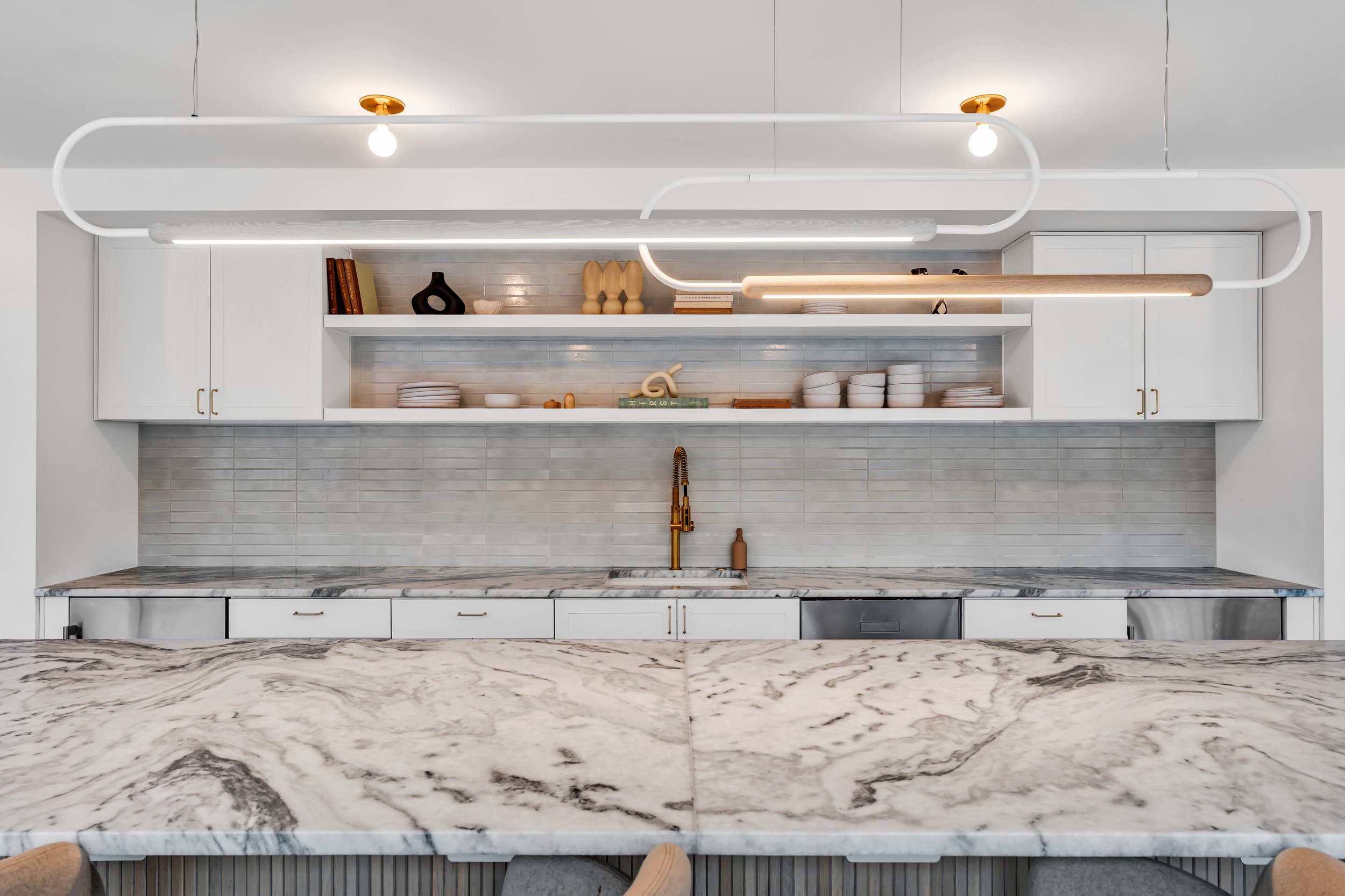 Modern kitchen with white cabinets, gray subway tile backsplash, and a marble countertop. Open shelves hold dishes, bowls, books, and decorative vases. A wooden and LED light fixture is suspended from the ceiling, and a gold faucet is visible over th