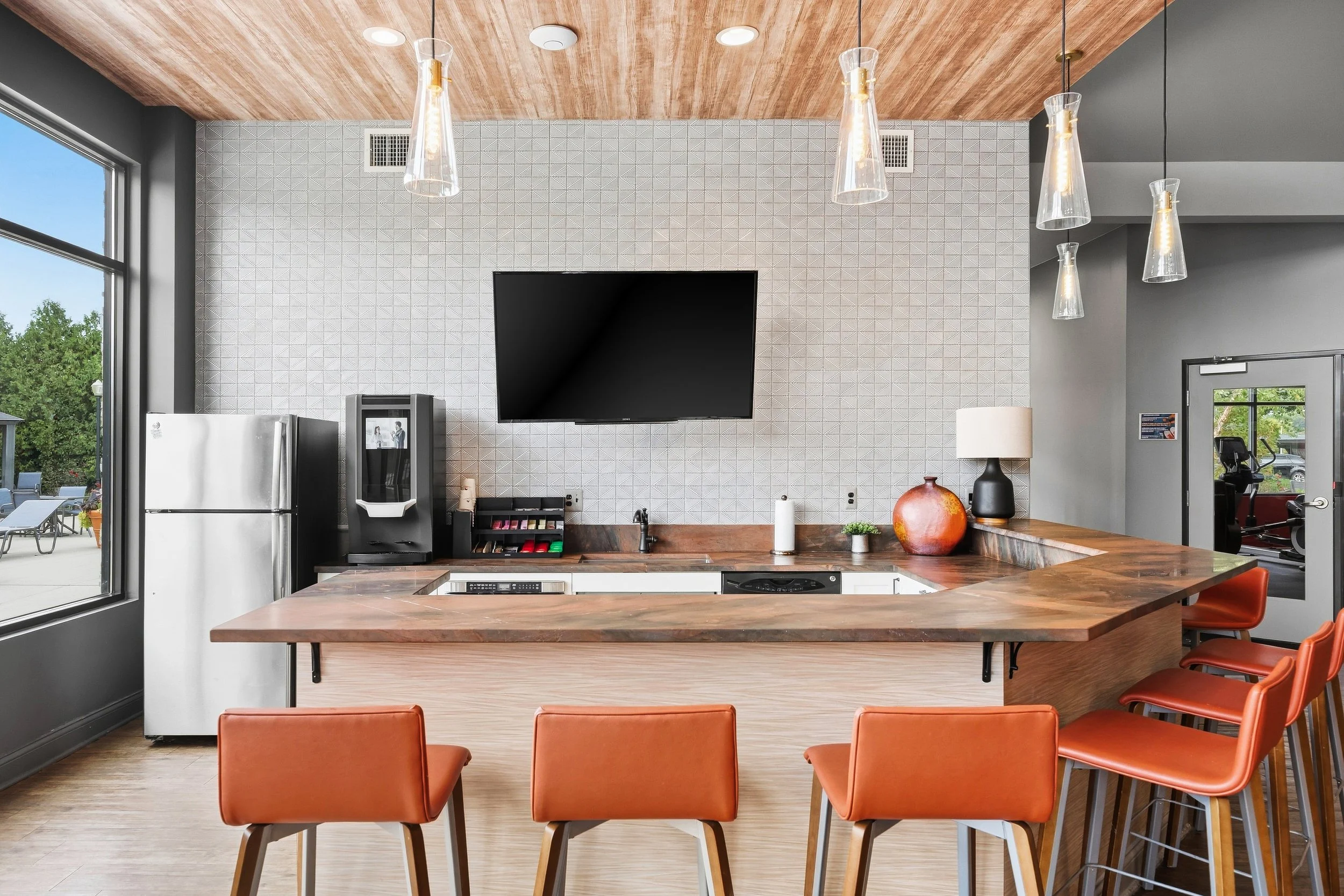 Modern kitchen with wooden island, orange chairs, gray walls, black microwave, stainless steel refrigerator, wall-mounted TV, glass pendant lights, and large windows showing outdoor trees.