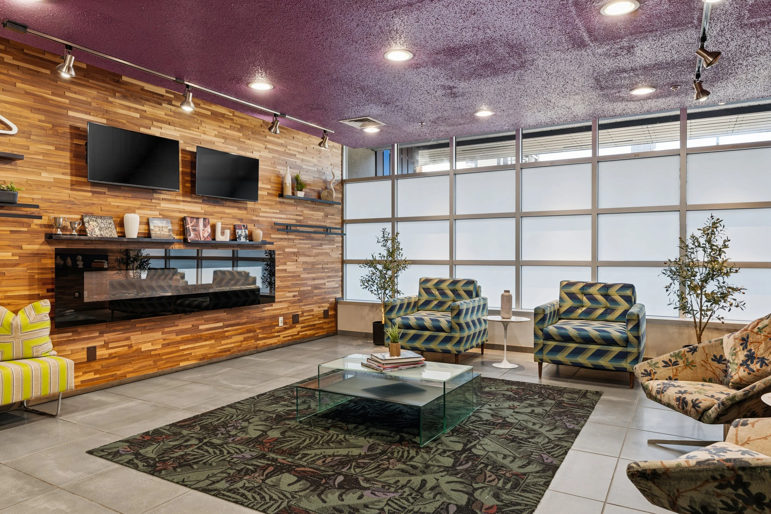 Modern lounge area with patterned armchairs, a glass coffee table, potted plants, a wood-paneled wall with mounted flat-screen TVs, decorative vases, and large windows with frosted glass panels.