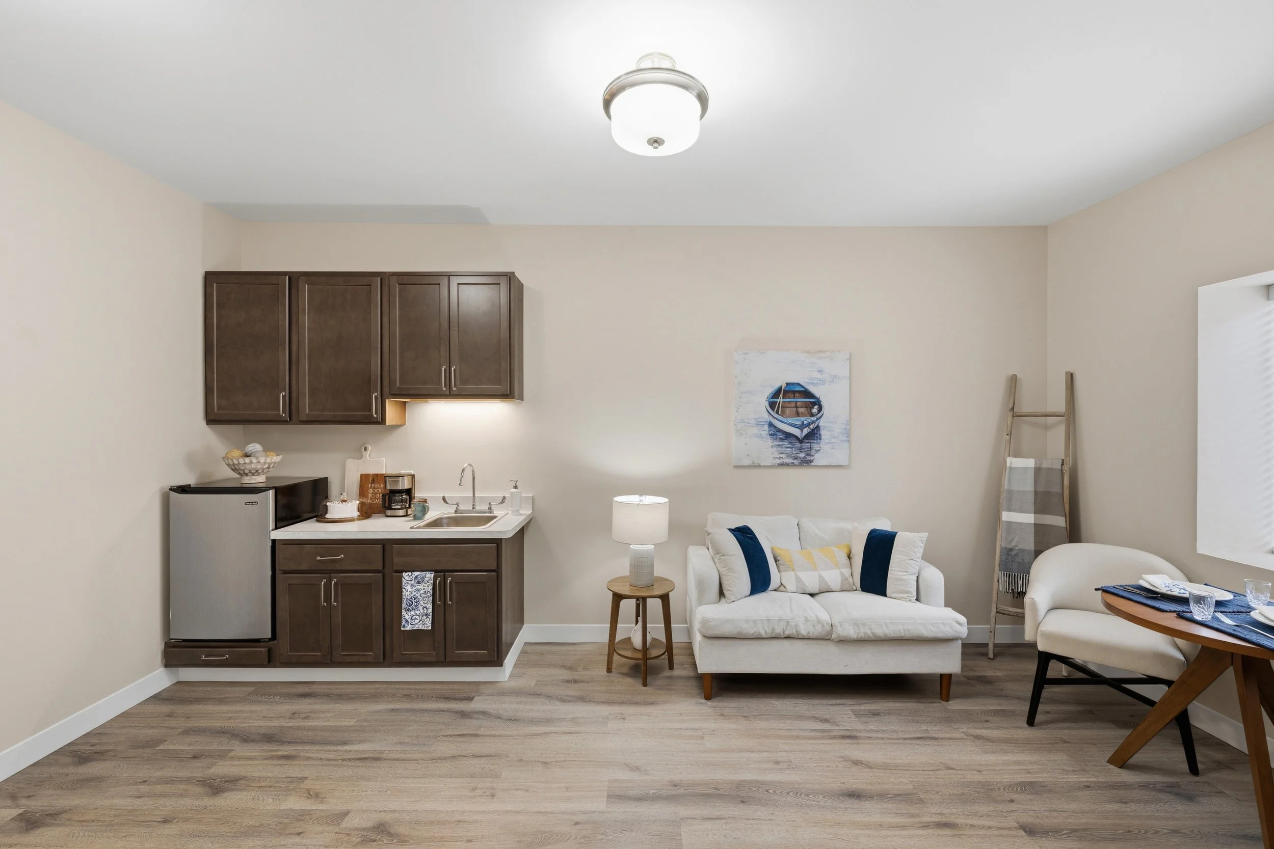 Interior view of a living room and kitchen area with beige walls and wood flooring; includes a white sofa with yellow and blue pillows, a small round side table with a lamp, a ladder with a striped blanket, and a painting of a boat on the wall.