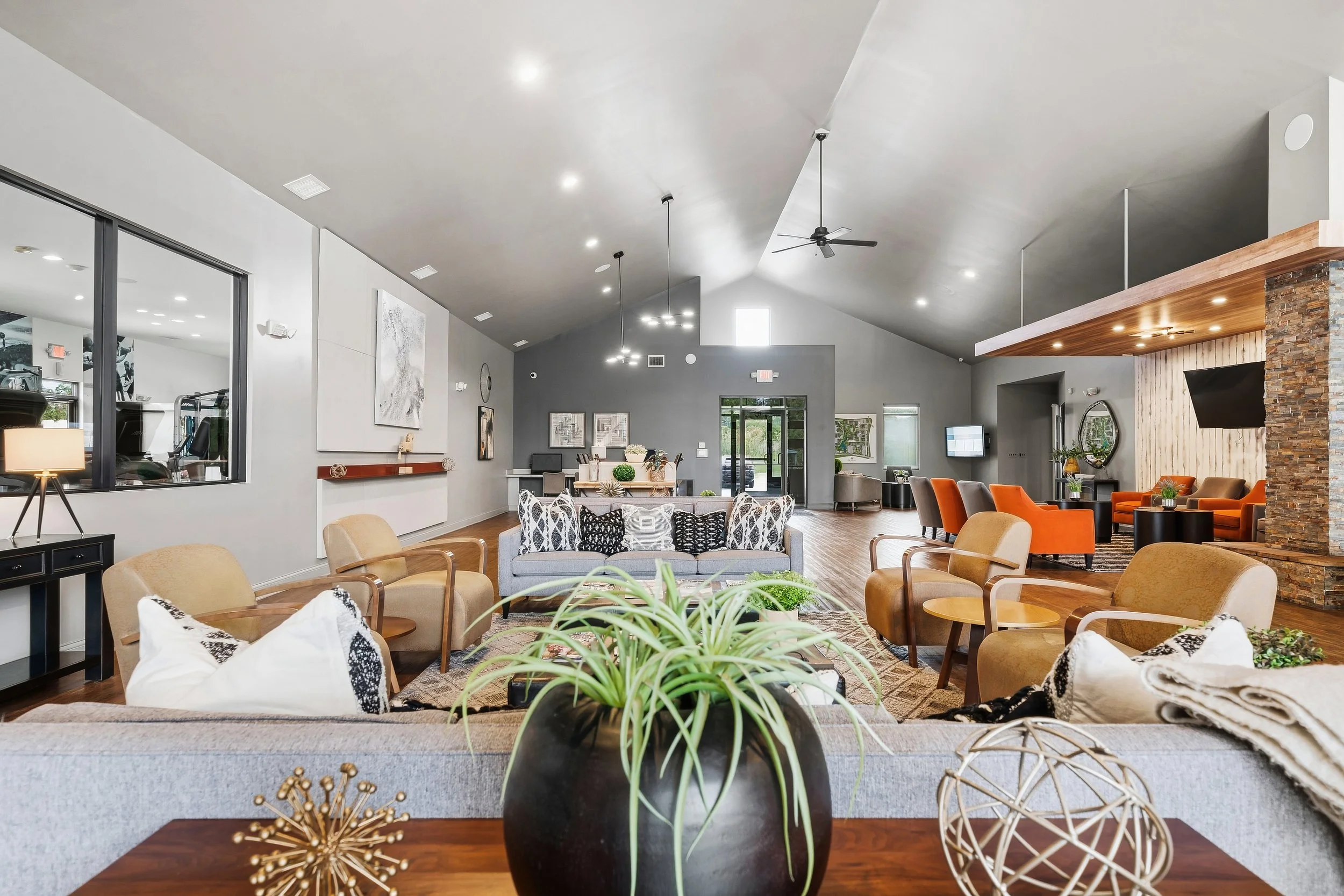 Spacious modern lounge with various seating including beige, gray, and orange chairs, and a wooden accent wall with a mounted flat-screen TV. Decorative plants, artwork, and contemporary decor are present. The room features high ceilings, large windo