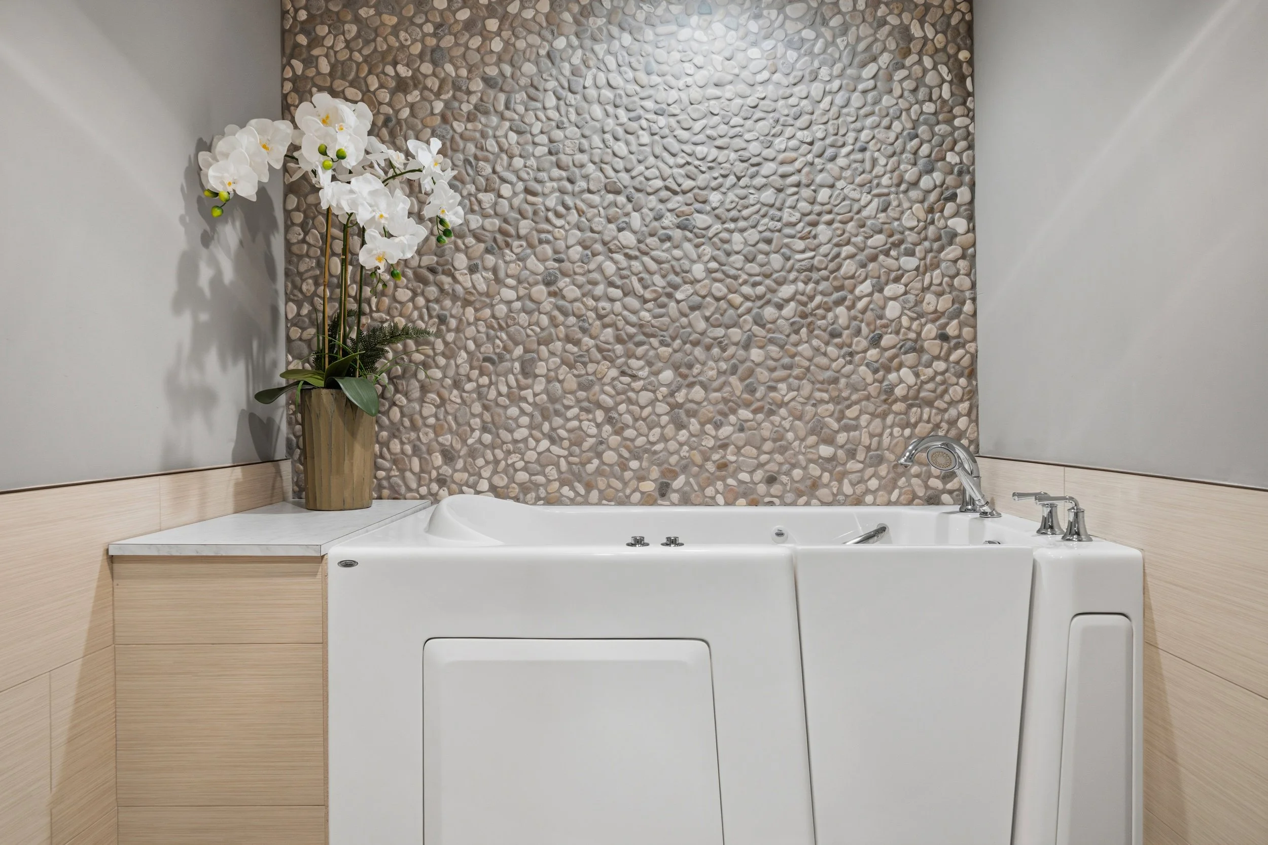 A bathroom with a white bathtub, a faucet, and controls, set against a wall with a pebble stone accent. There is a potted orchid with white flowers on a ledge beside the tub.