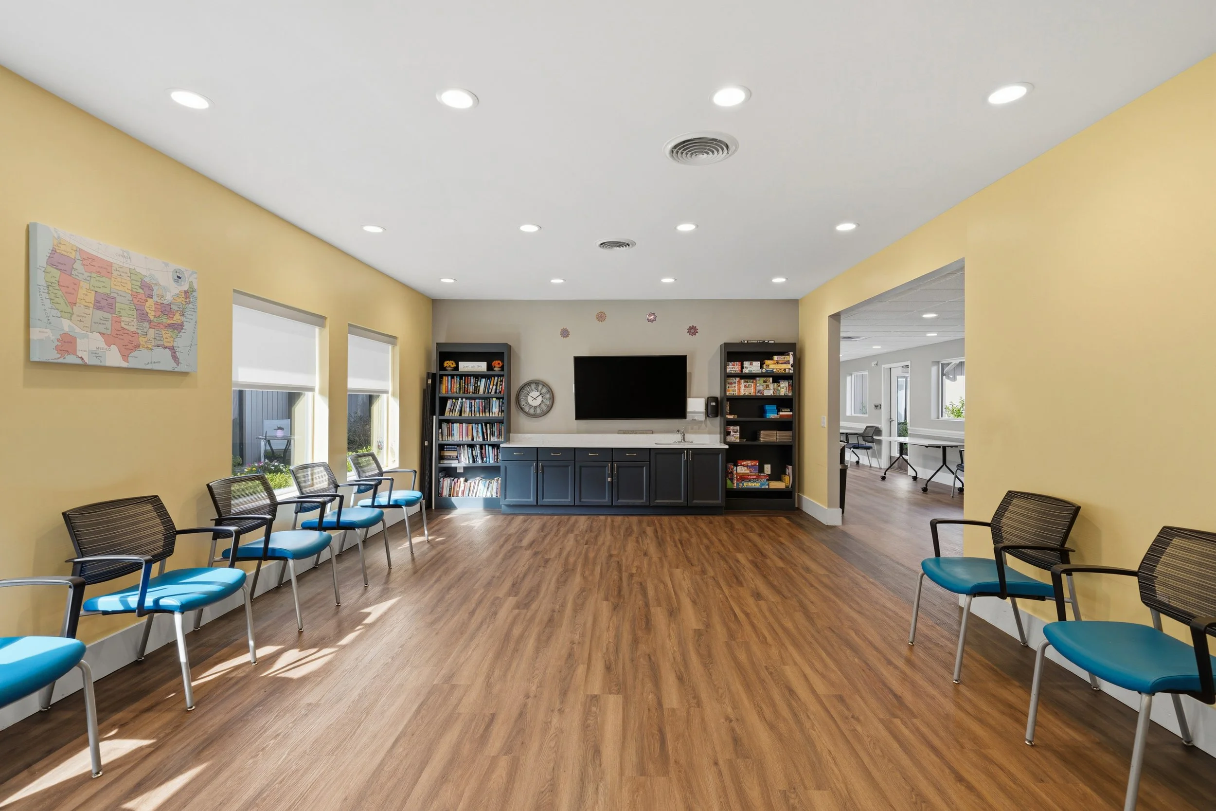 A waiting room with two rows of blue chairs with black armrests along a yellow wall. A large map of the United States is on the wall. A door and windows are visible, with a TV, bookshelf, and wall clock in the background.