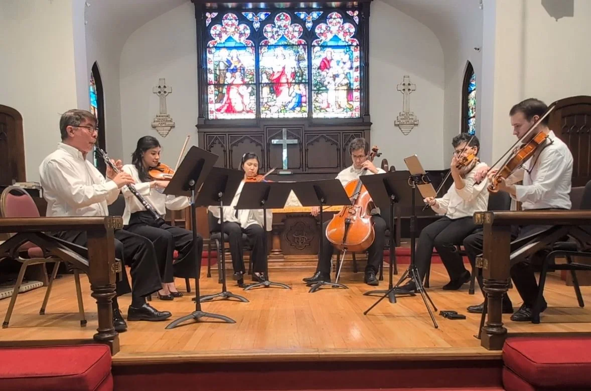 A group of six musicians performing on stage in a church with stained glass windows in the background. They are playing various instruments, including oboe, violin, cello, and viola, and are dressed in formal white shirts.