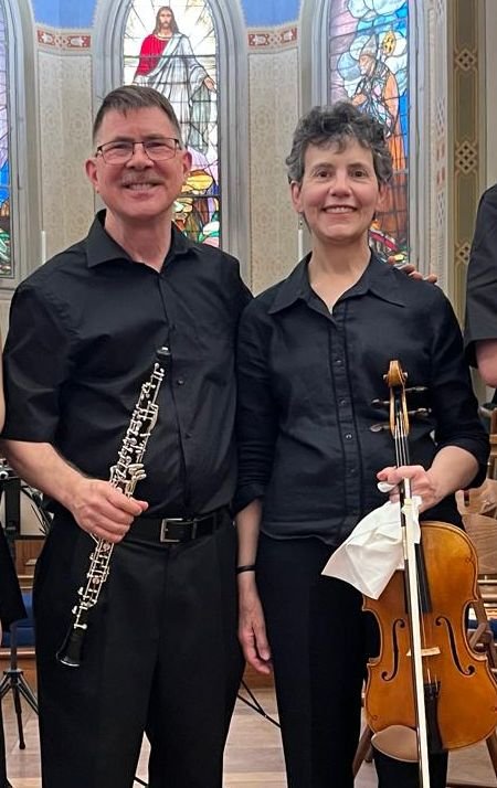Two musicians, a man and a woman, standing inside a church with stained glass windows, holding a clarinet and a violin, both smiling and dressed in black.