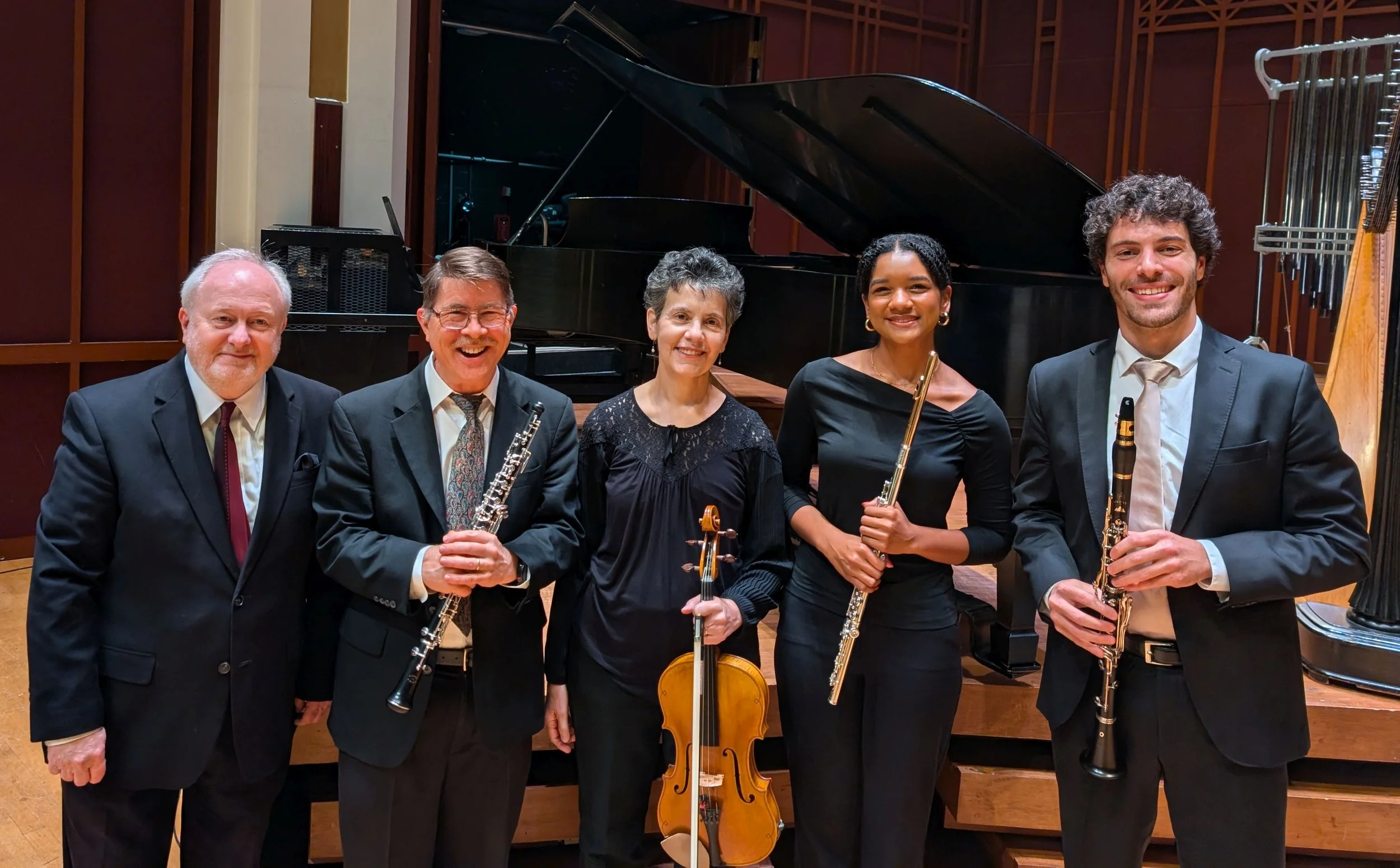 Five musicians posing with their instruments in a concert hall, with a grand piano in the background.