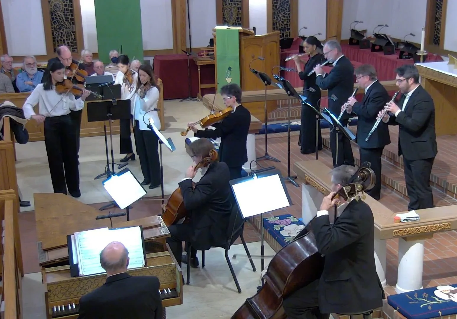 An orchestral performance inside a church or concert hall with musicians playing violins, flutes, clarinets, and cellos, and an audience seated in the background.