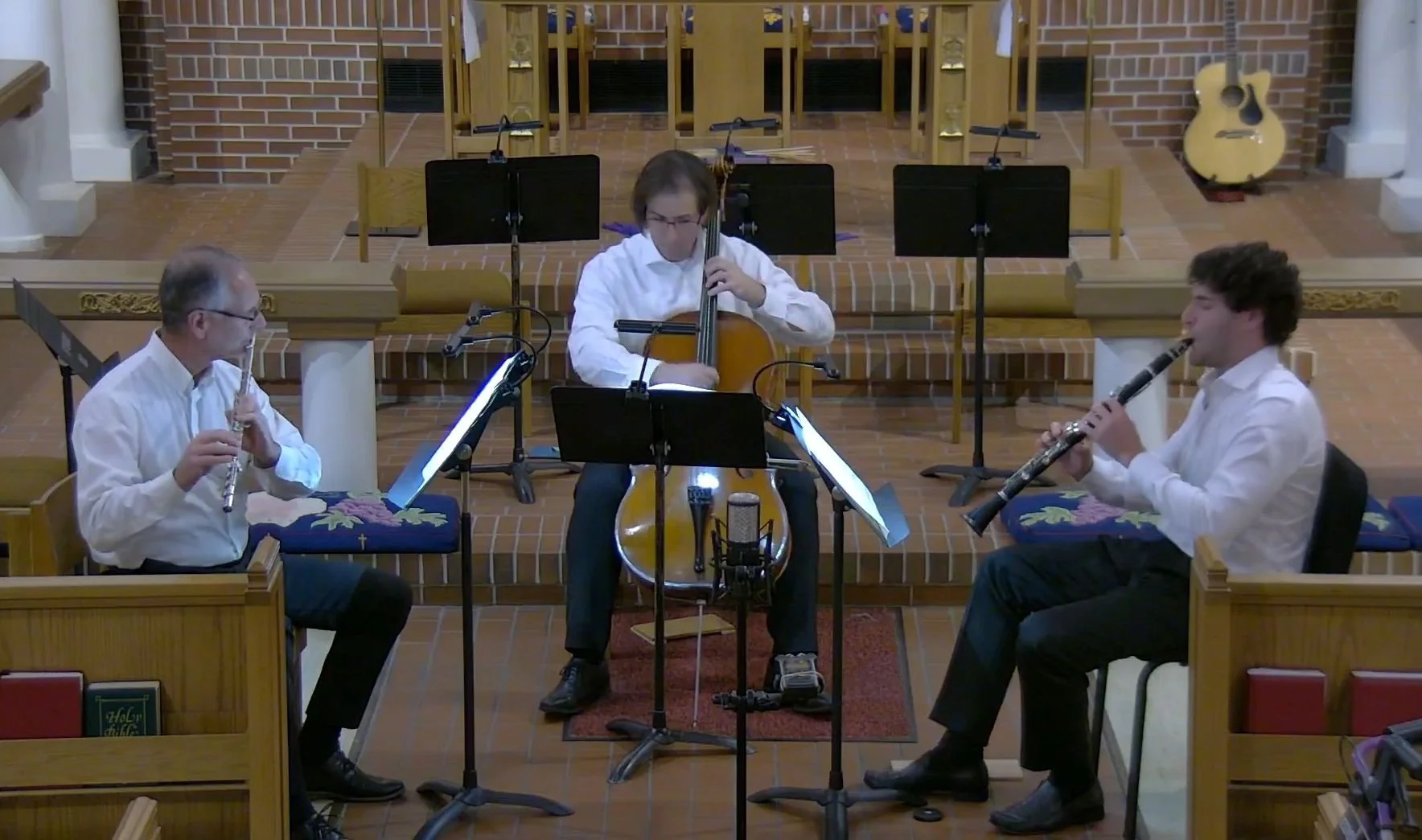 Three musicians playing during a performance in a church. The musicians include a man playing a flute on the left, a man playing a cello in the center, and a man playing a clarinet on the right. They are seated in front of music stands with sheet music. The setting has brick walls, wooden pews, and a guitar leaning against a brick wall in the background.