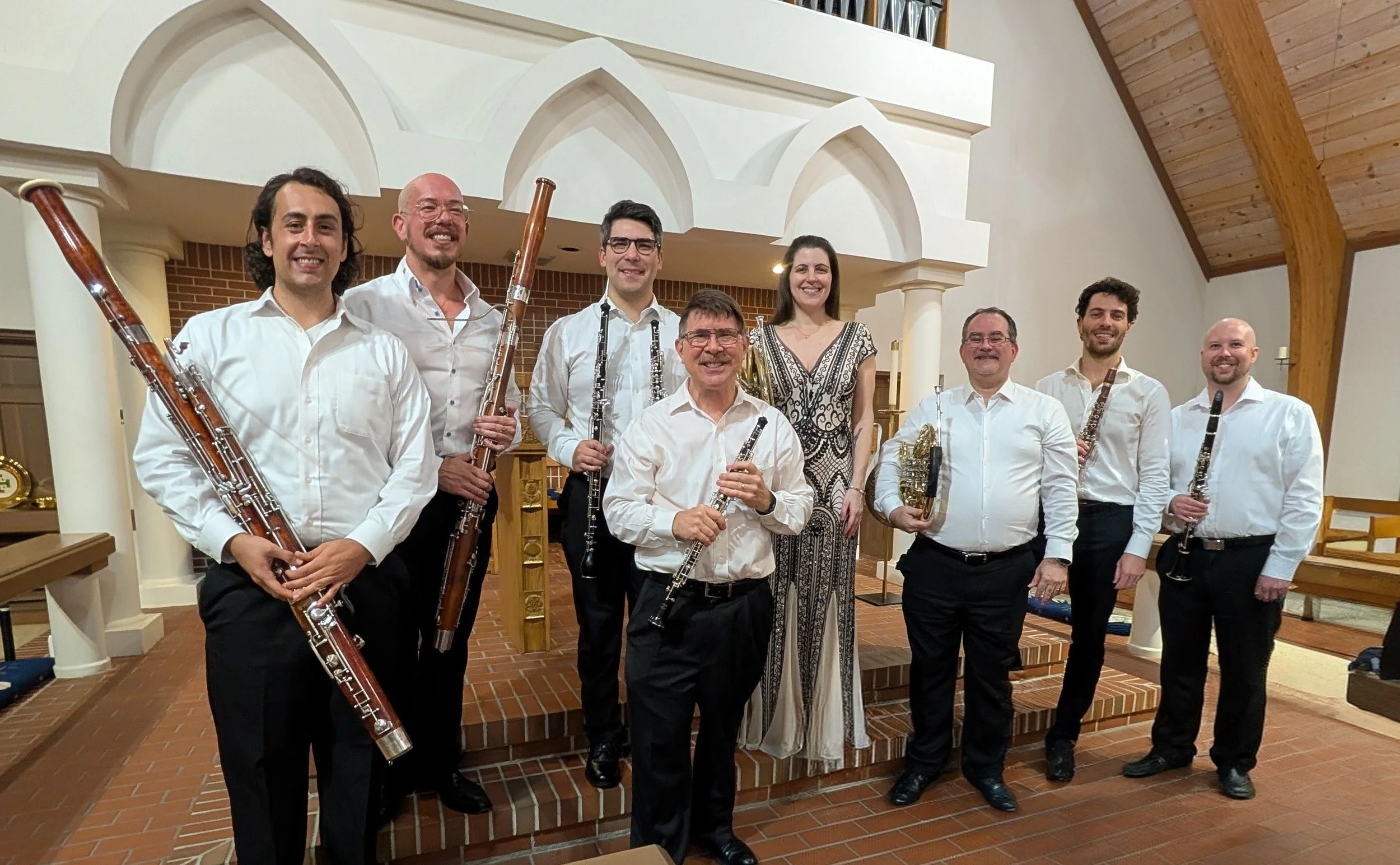 Group of nine musicians with musical instruments posing inside a church or concert hall, smiling for the camera.