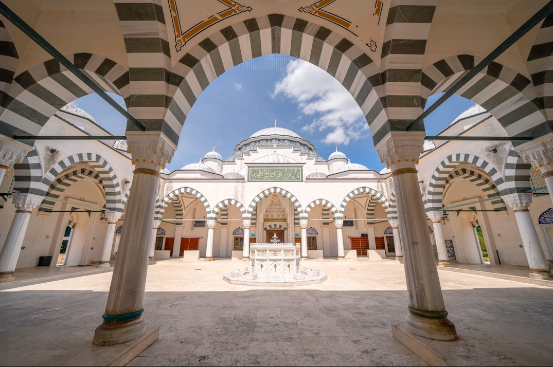 Interior courtyard of a mosque with striped arches, domes, and a central fountain, under a blue sky with clouds.