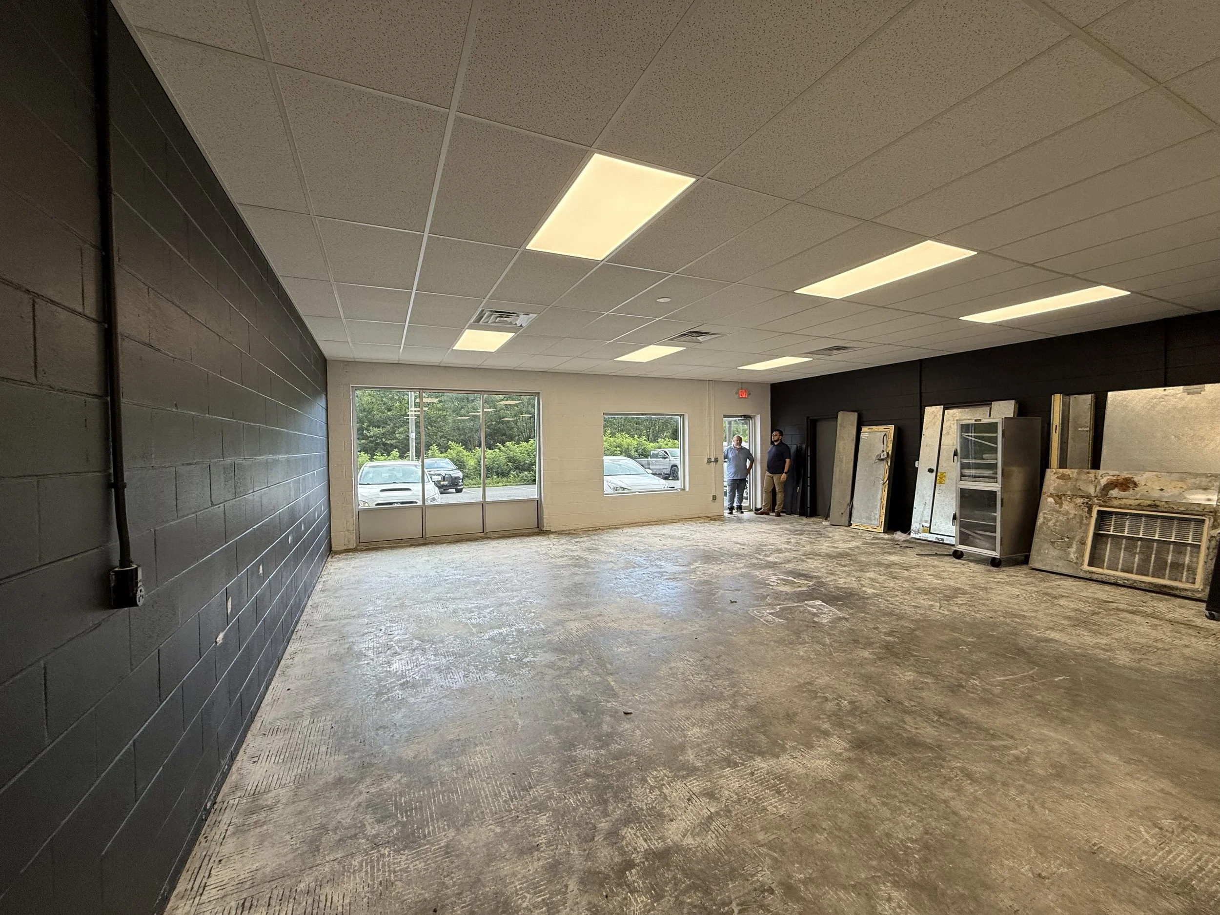 Interior of a commercial space with unfinished concrete flooring, black brick walls, and ceiling tiles with built-in fluorescent lights. Two people are standing near the back, with large windows and doors revealing a parking lot and greenery outside.