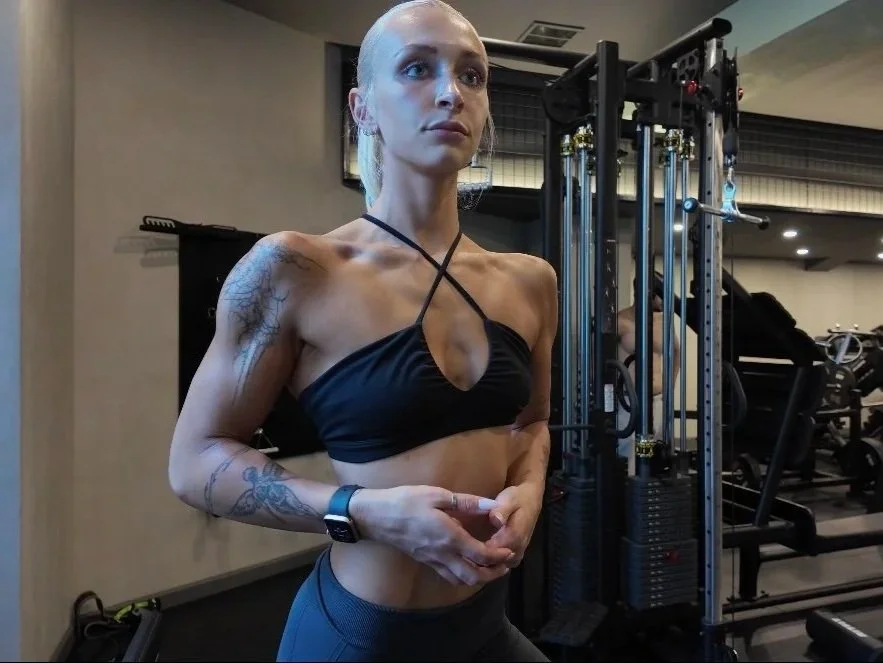 A woman at the gym stands in front of gym equipment wearing a black sports bra with a crisscross strap design and dark leggings, with tattoos on her left arm and a fitness tracker on her right wrist.
