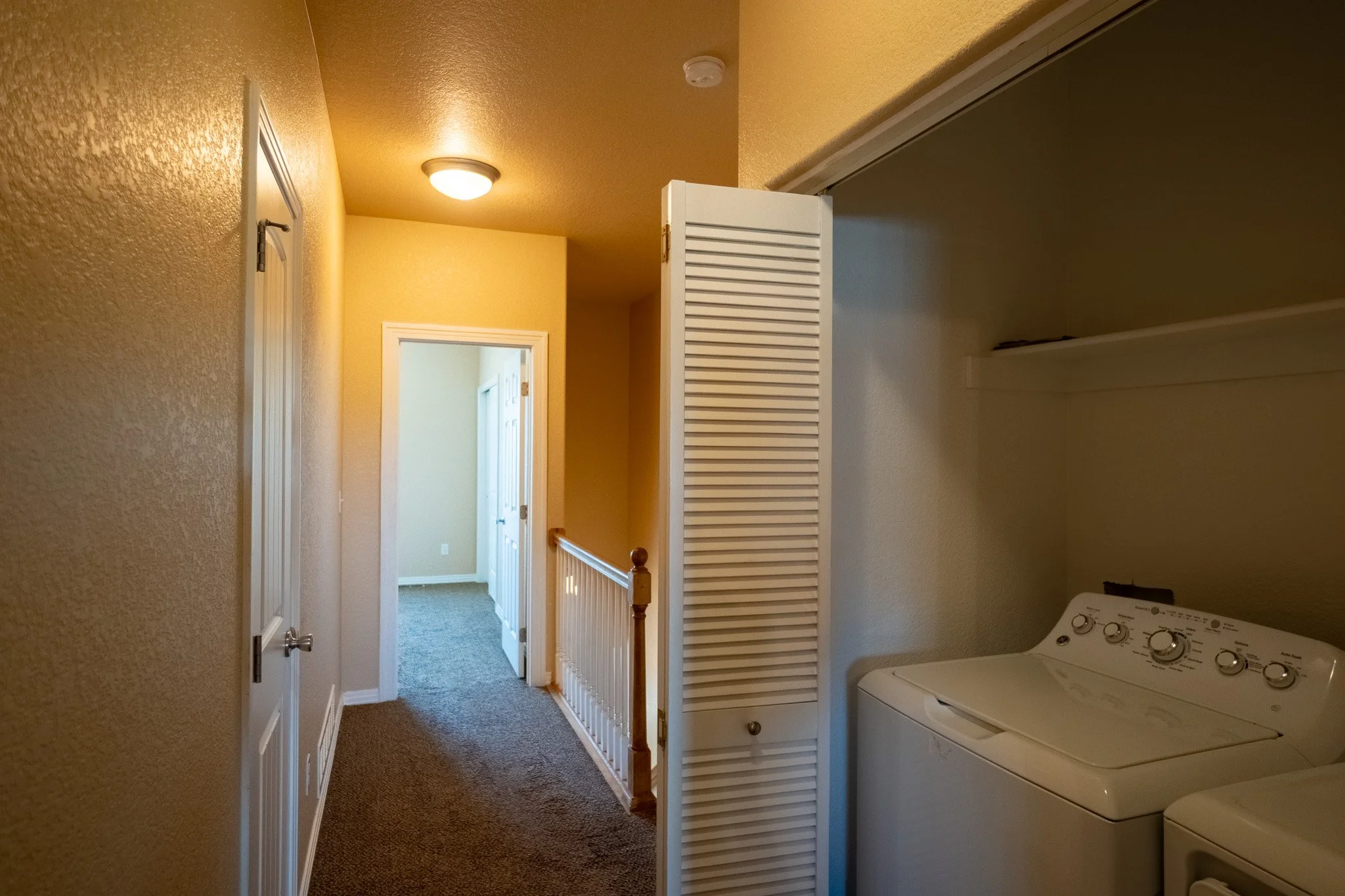 A laundry room with a white washing machine and a vented door partition, beige walls, carpeted floor, and ceiling light fixture.