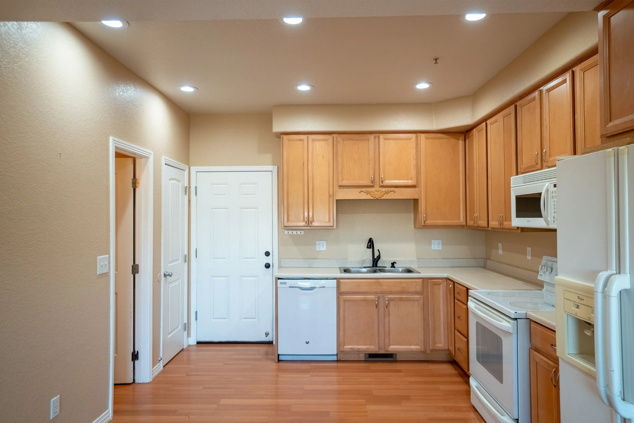 View of a kitchen with wooden cabinets, white appliances including a stove and microwave, a black faucet, beige countertops, and beige walls with a textured finish. The ceiling has recessed lighting.