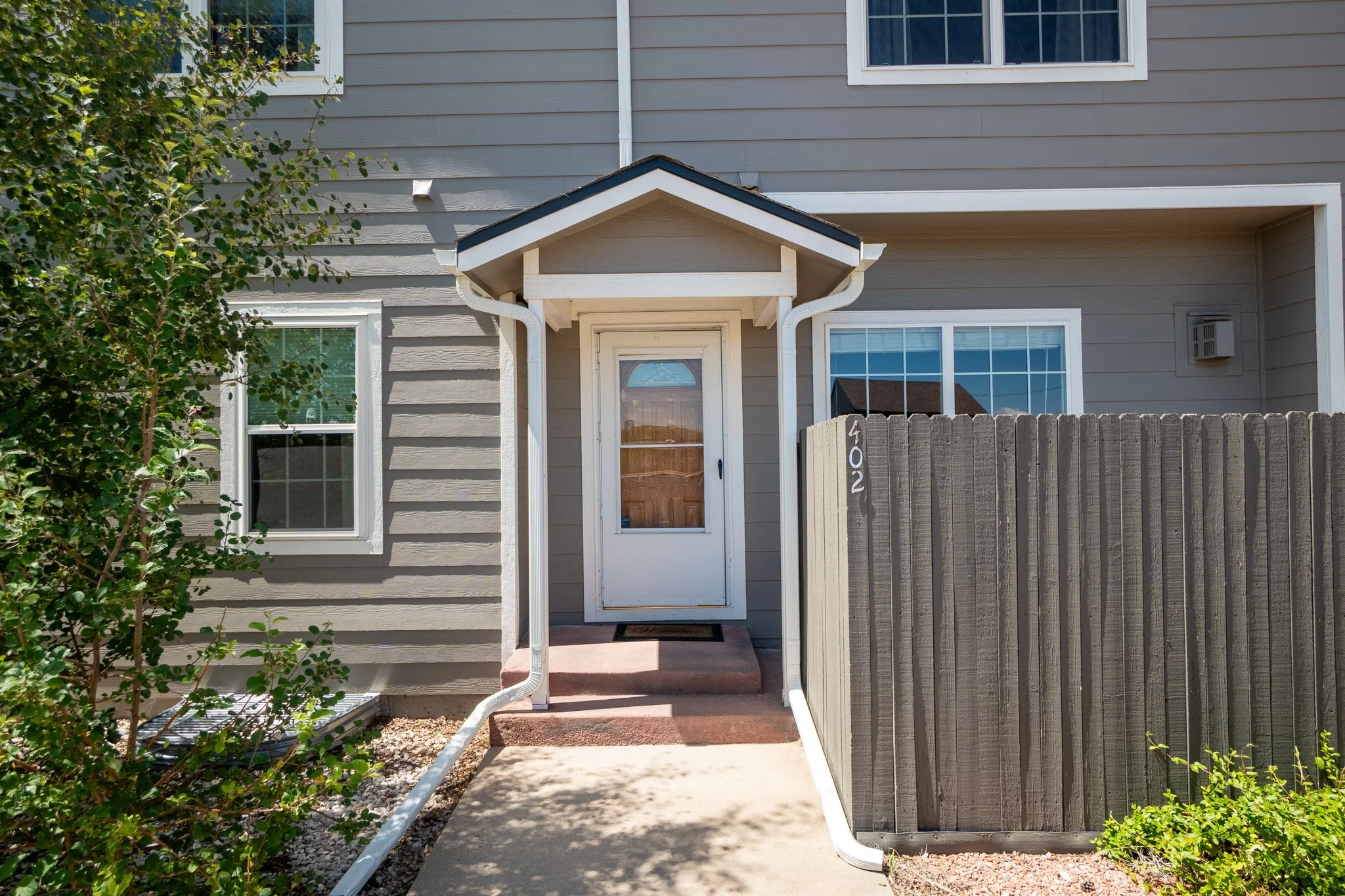 Front entrance of a beige house with a small covered porch, white door, and a large window. There is a wooden fence on the right and a small tree on the left.