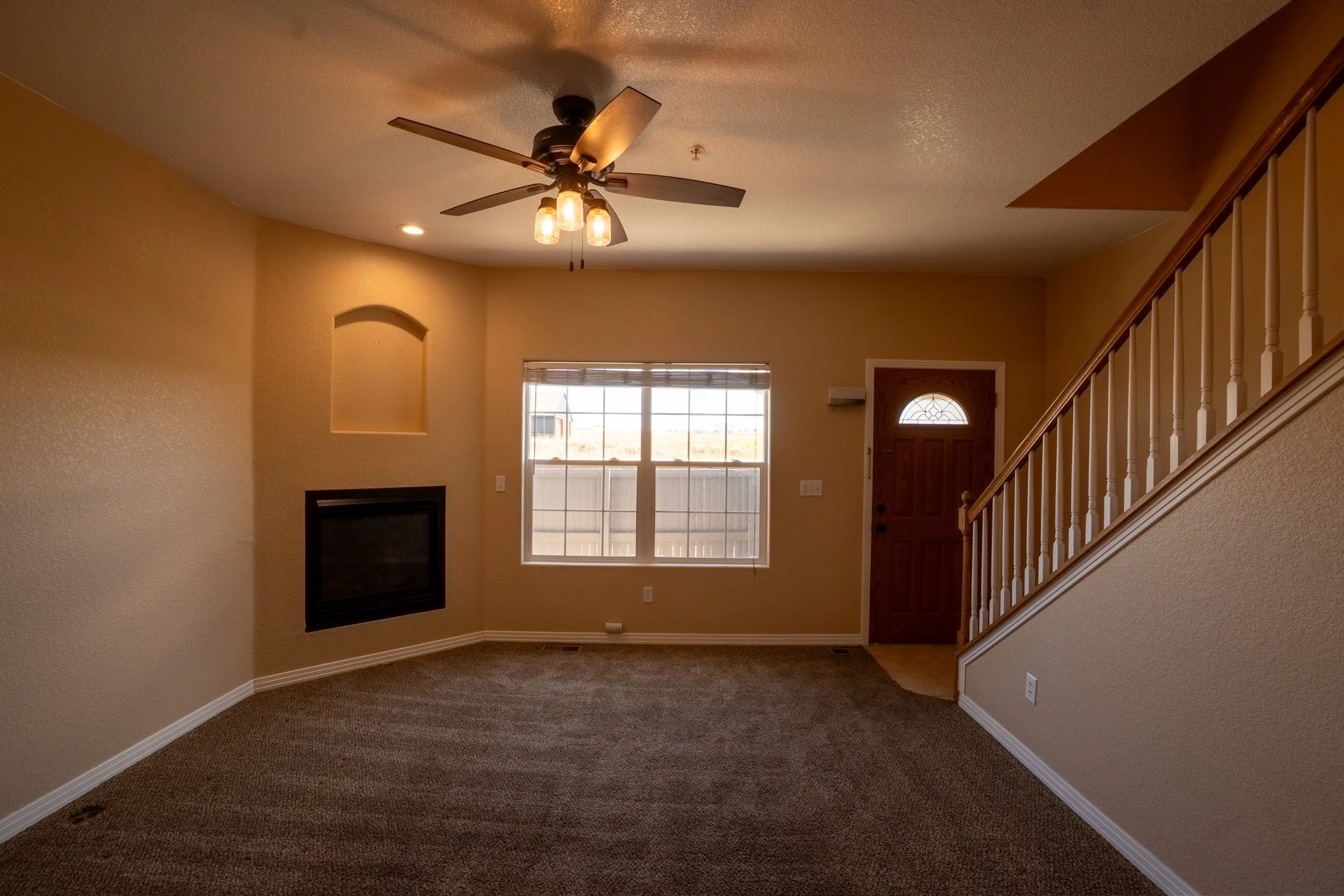 Empty living room with beige walls, a window, a door, a staircase, a ceiling fan with lights, and a built-in wall niche.