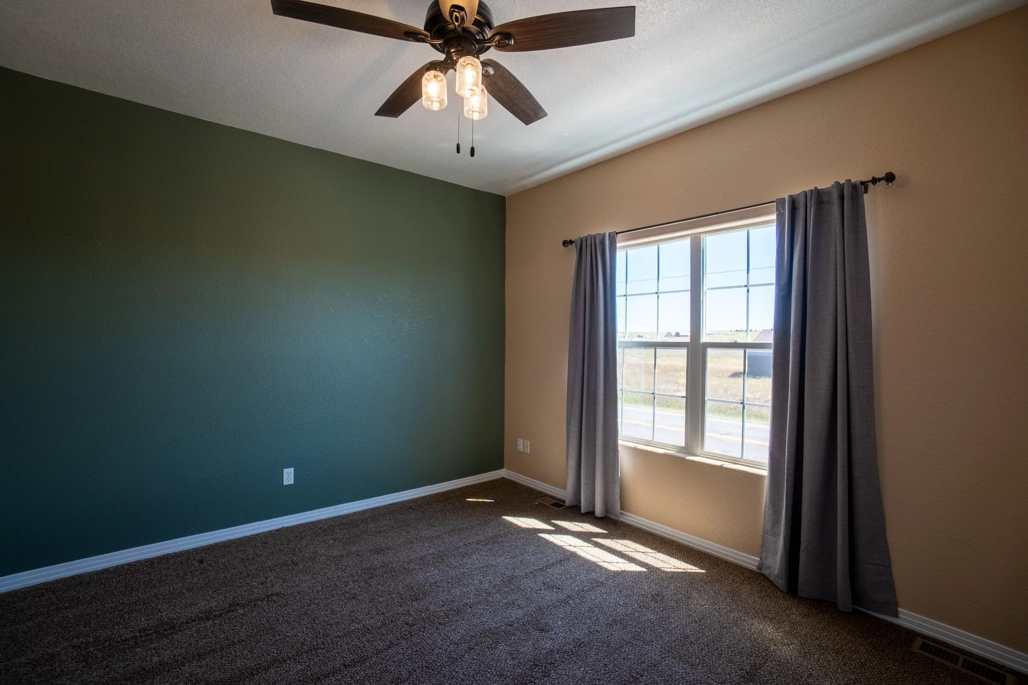 Empty room with one green wall and one beige wall, brown carpet, a window with gray curtains, sunlight streaming in, and a ceiling fan with lights.