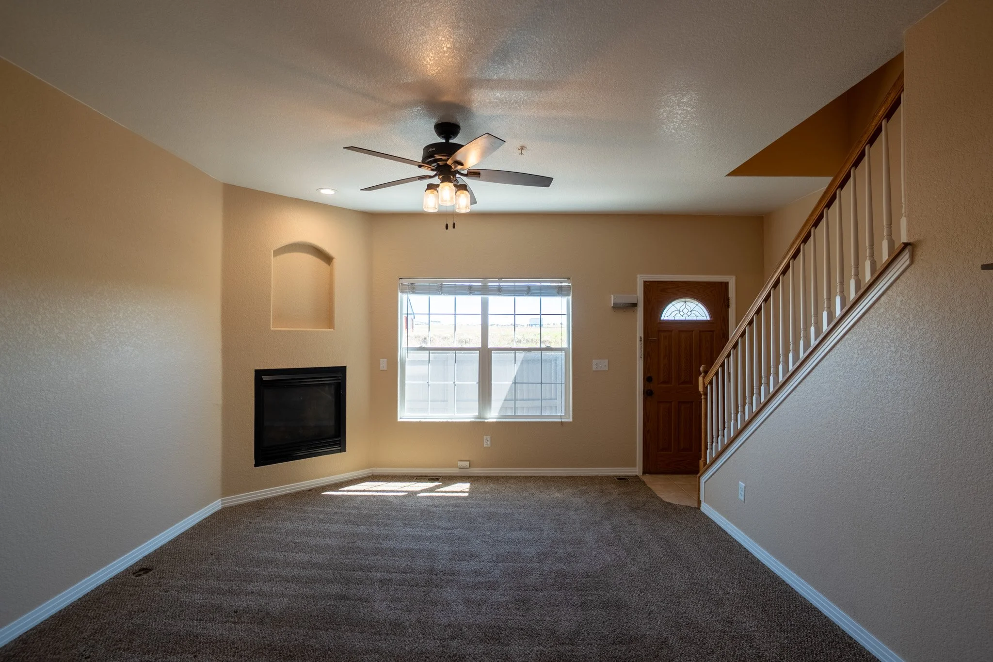 Empty living room with beige walls, a ceiling fan, a window, a staircase, and a fireplace.